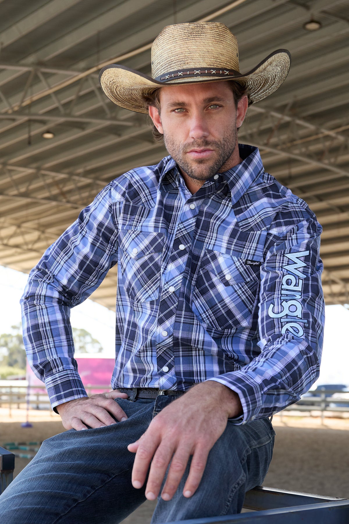 Wrangler Men's Billy Logo L/S Western Shirt. Man wearing a blue plaid shirt with 'Wrangler' branding and a cowboy hat indoors.