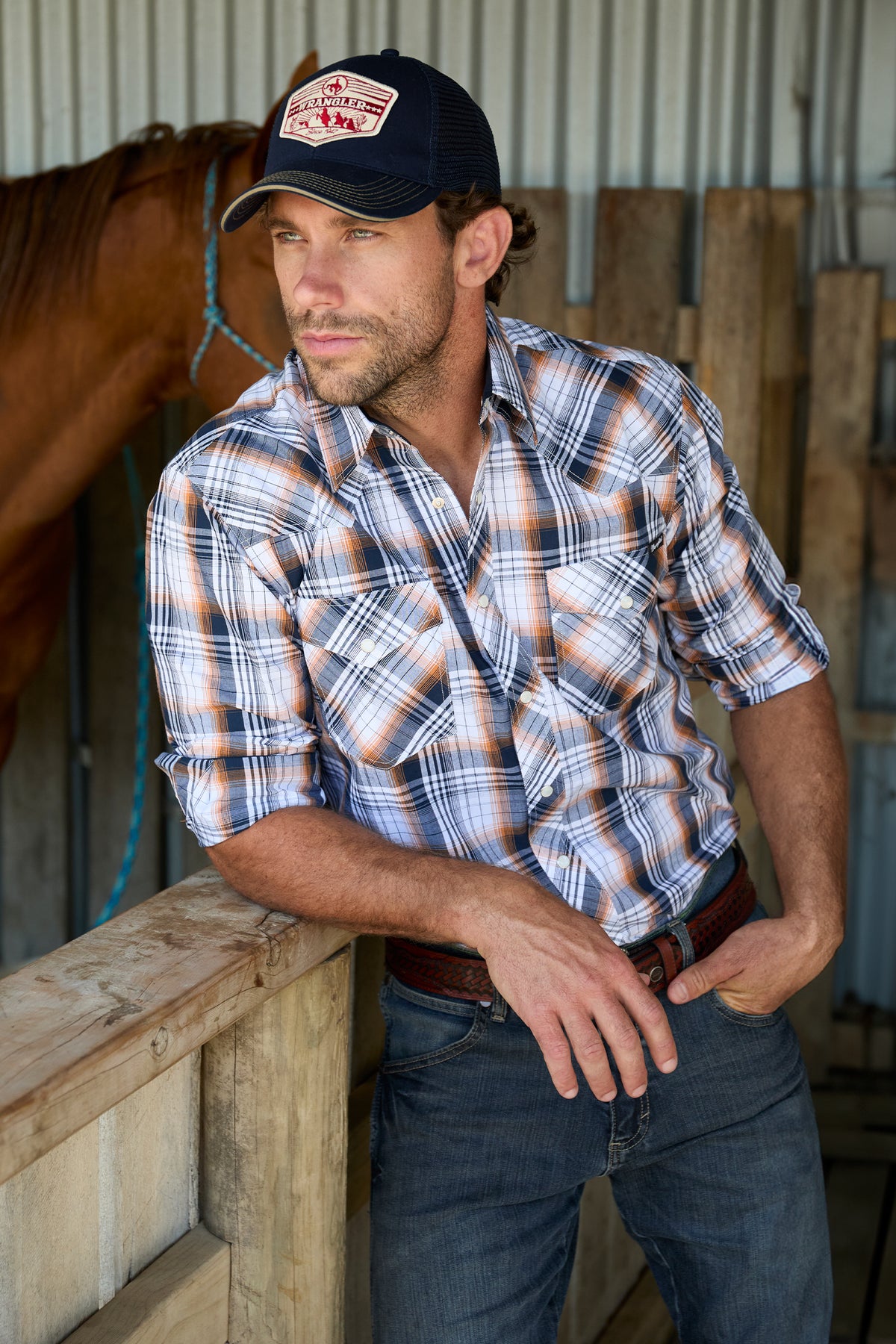 Wrangler Men's Toby L/S Western Shirt. Man wearing a plaid shirt and cap standing next to a horse in a barn.