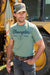 Wrangler Men's Arthur S/S T-Shirt. Man wearing a Wrangler t-shirt and cap standing in front of a yellow piece of machinery.
