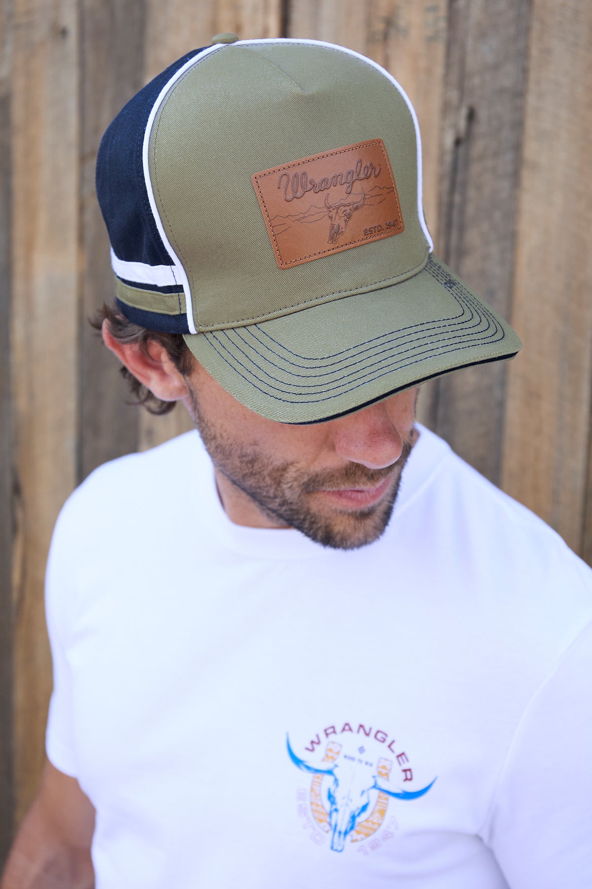 Wrangler Men's Damon High Profile Cap. Man wearing a green and navy cap with a leather patch, standing against a wooden fence.