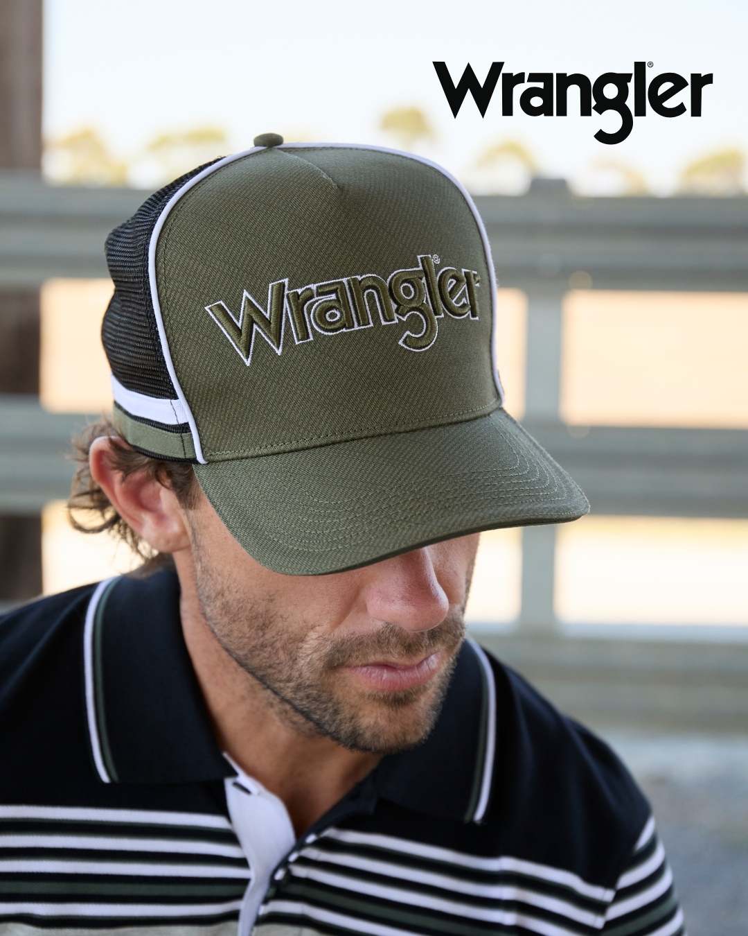 Wrangler Men's Logo High Profile Trucker Cap. Man wearing a green cap with 'Wrangler' logo, blurred background