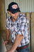 Wrangler Men's Trevor 6 Panel Trucker Cap. Man wearing a plaid shirt and cap with a logo, standing against a wooden background
