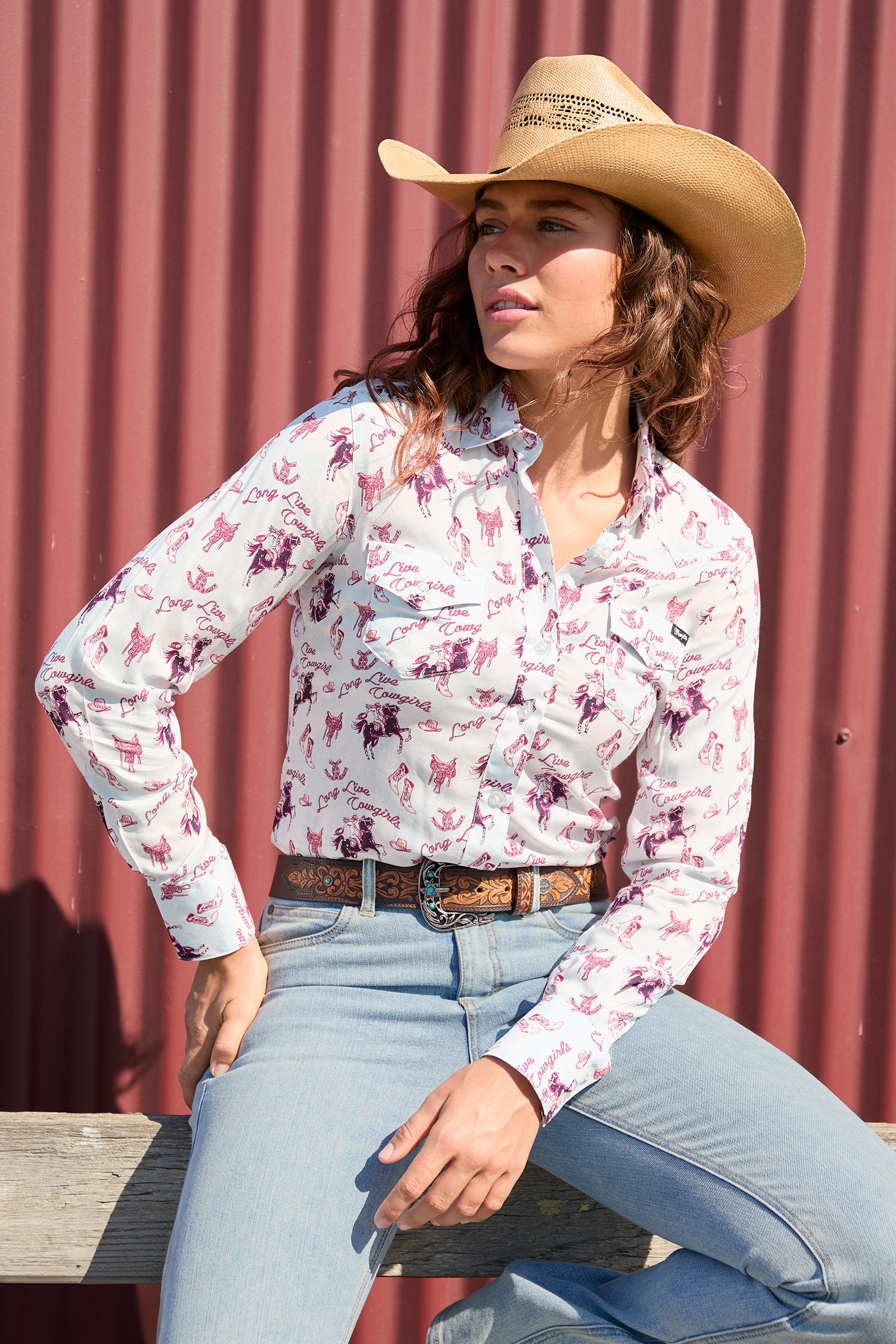 Wrangler Women's Doreen L/S Western Shirt. Woman wearing a floral shirt, jeans, and a cowboy hat against a red corrugated metal background.
