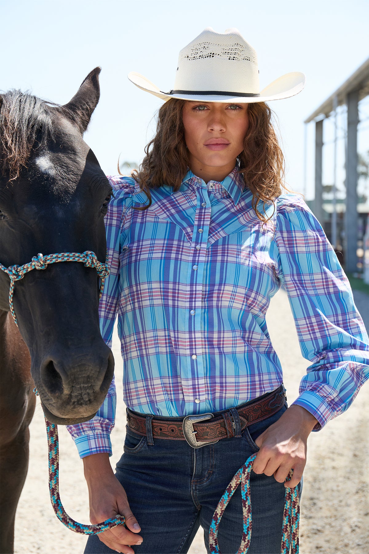 Wrangler Women's Emily Check L/S Western Shirt. Person wearing a blue plaid shirt and white cowboy hat standing next to a horse.