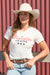 Wrangler Women's Piper S/S T-Shirt. Woman wearing a white 'Wrangler Jeans' t-shirt and cowboy hat against a red corrugated metal background.