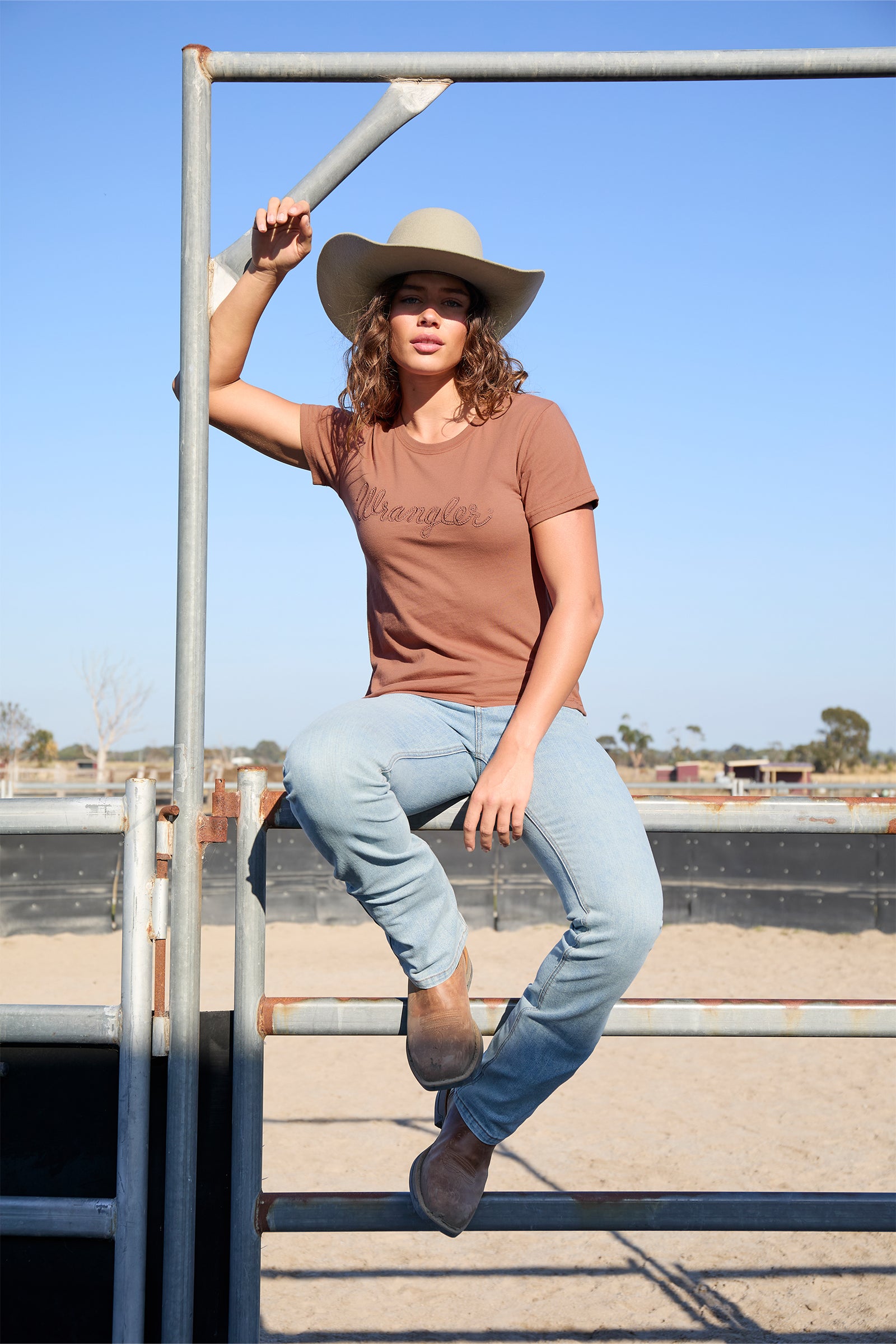 Wrangler Women's Texas S/S T-Shirt. Woman wearing a brown t-shirt, light blue jeans, and a beige hat, sitting on a metal railing.