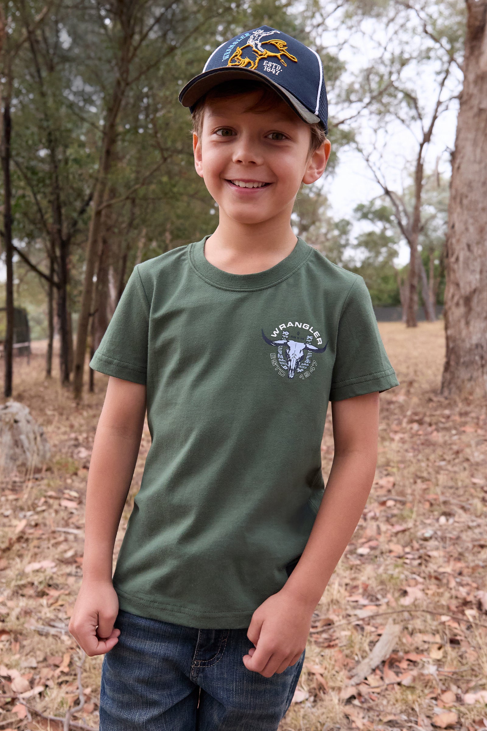 Wrangler Boy's Henry S/S T-Shirt. Child wearing a green t-shirt with a logo and a cap, standing in a forested area.