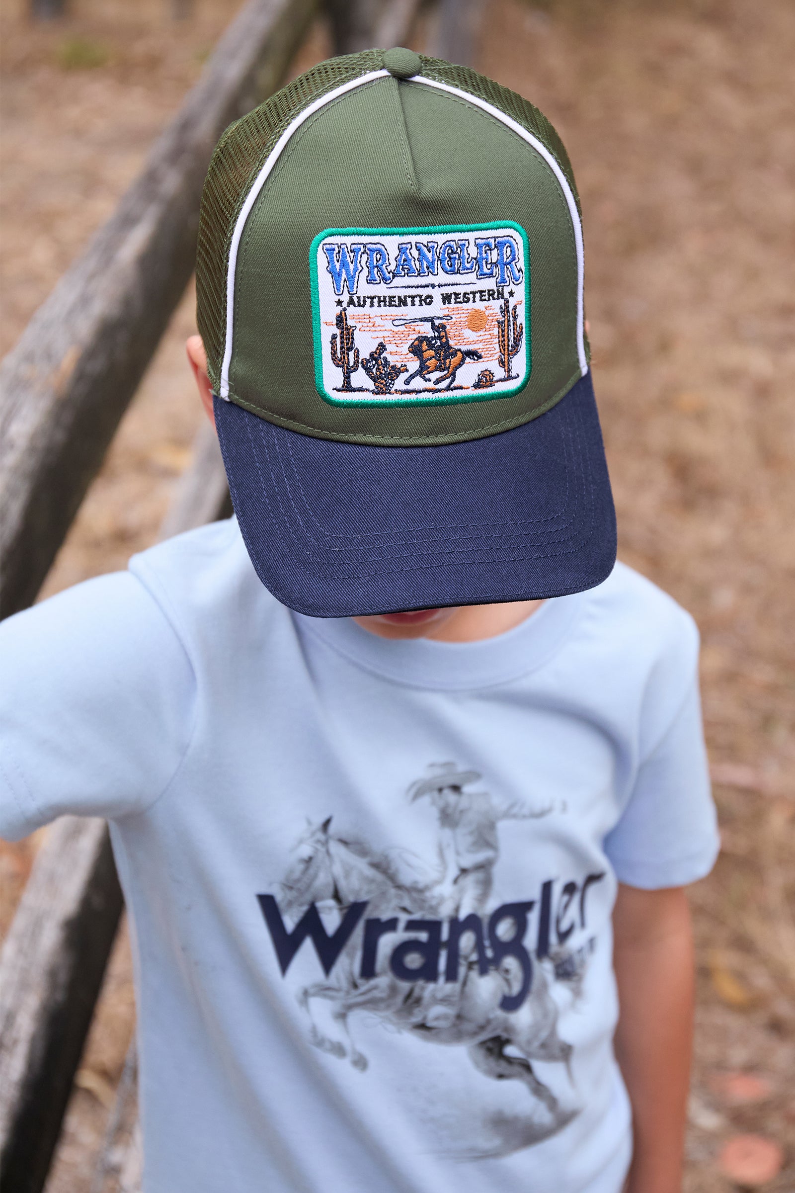 Wrangler Boy's Andy Trucker Cap. Child wearing a green and navy cap with Wrangler logo, standing outdoors.