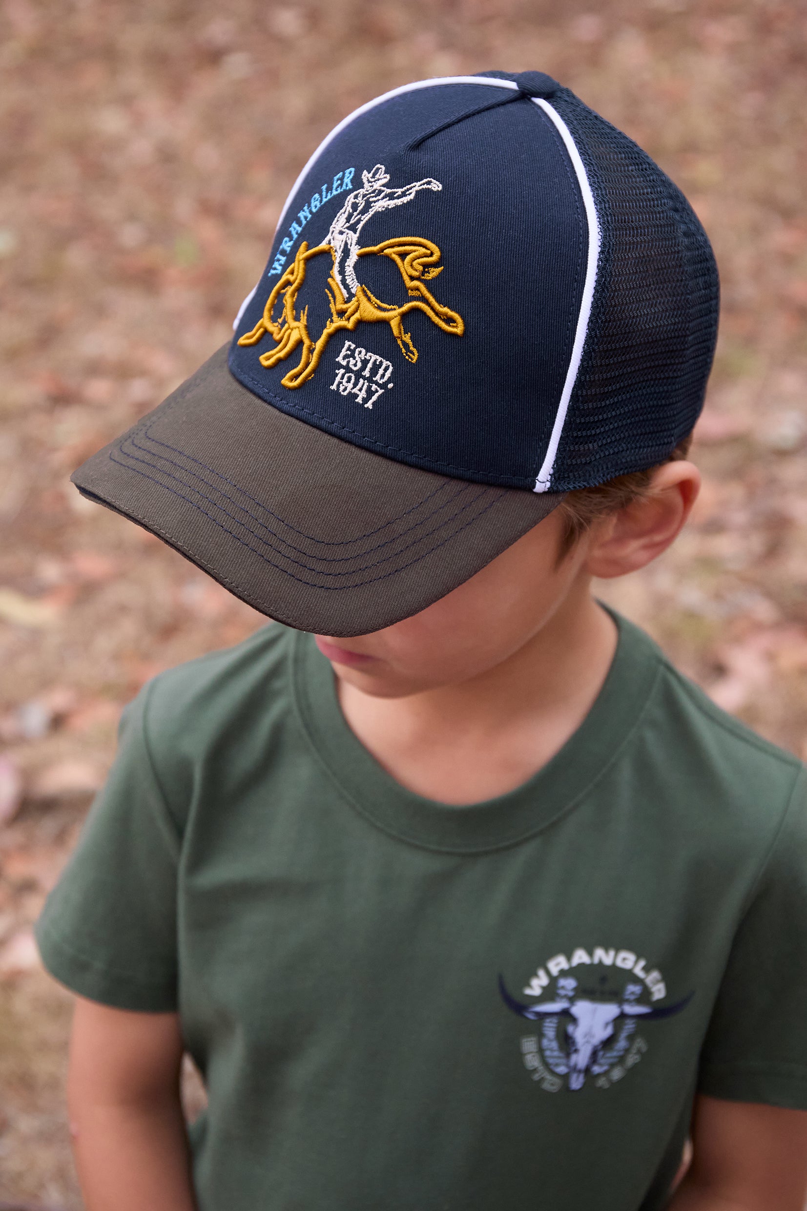 Wrangler Boy's Robert Trucker Cap. Child wearing a navy and brown cap with a logo, standing against a natural background.