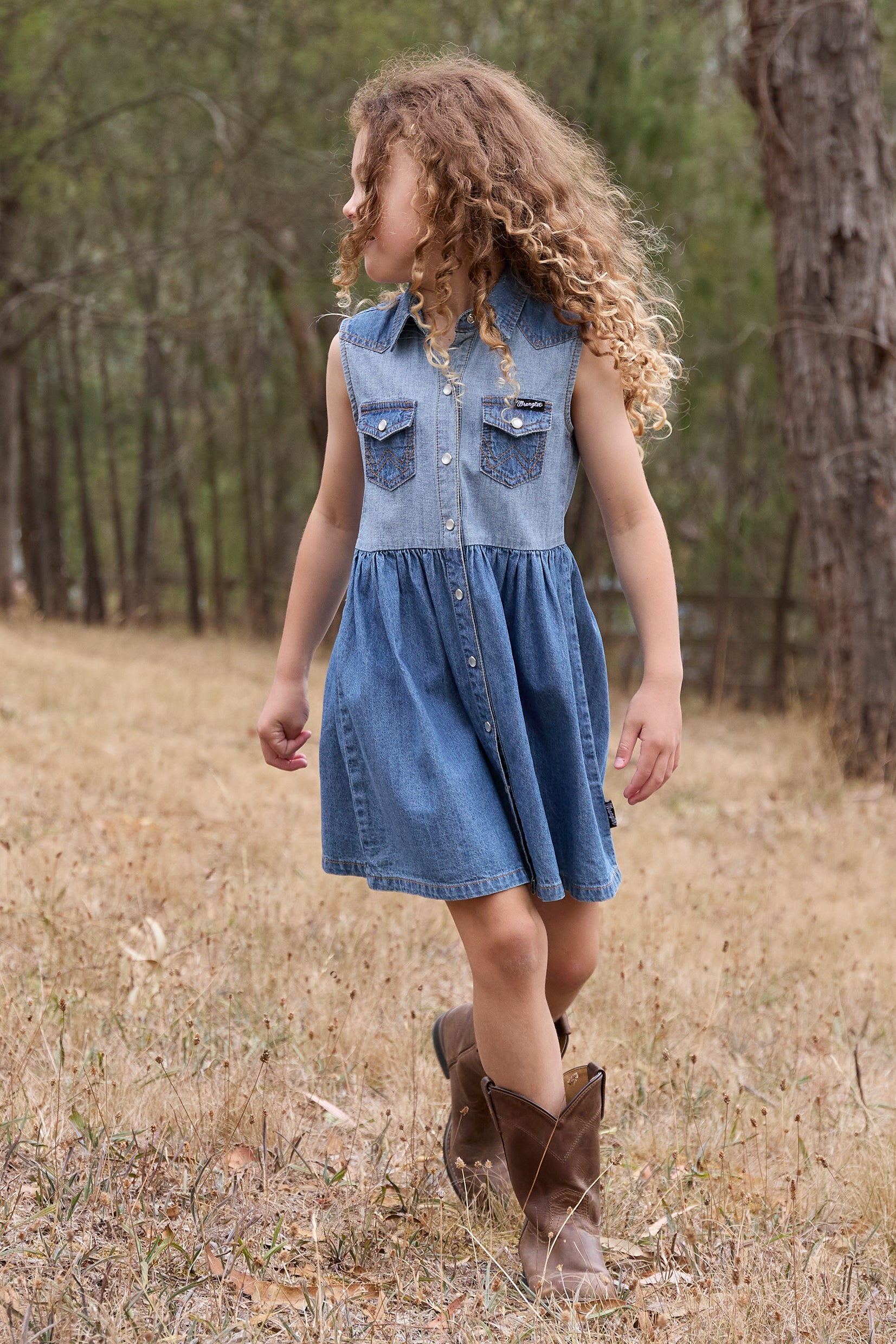 Wrangler Girl's Joanna Sleeveless Dress. Young girl in a denim dress and brown boots standing in a forested area.
