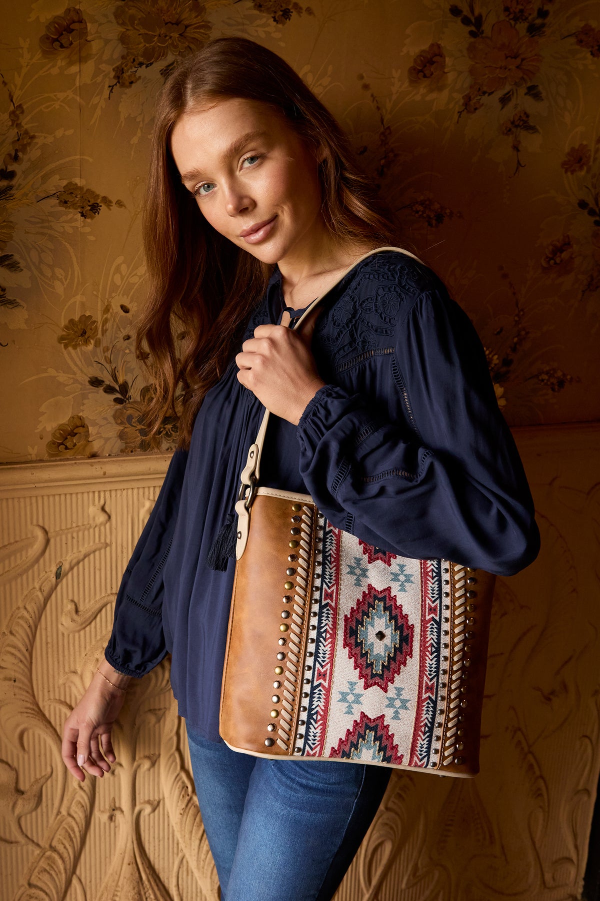 A brown Wrangler Women's Carmen Western Bag with a geometric pattern on the front, featuring a shoulder strap, metal feet, and a top zip opening.