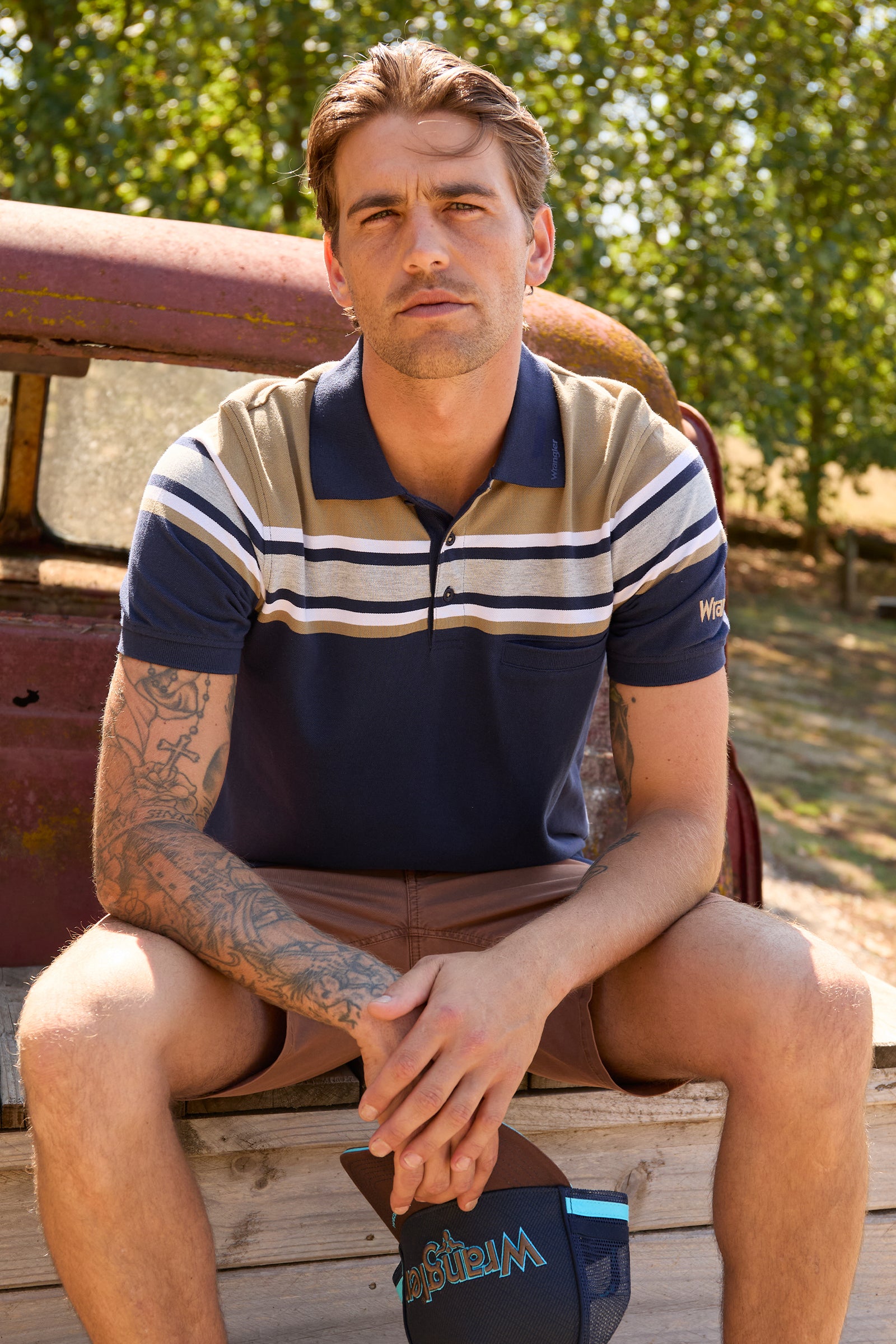 A man wearing a navy and beige striped polo shirt sits on the wooden bed of a vintage pickup truck outdoors. A navy blue baseball cap with 'Miz Mooz' written on it rests on his lap.