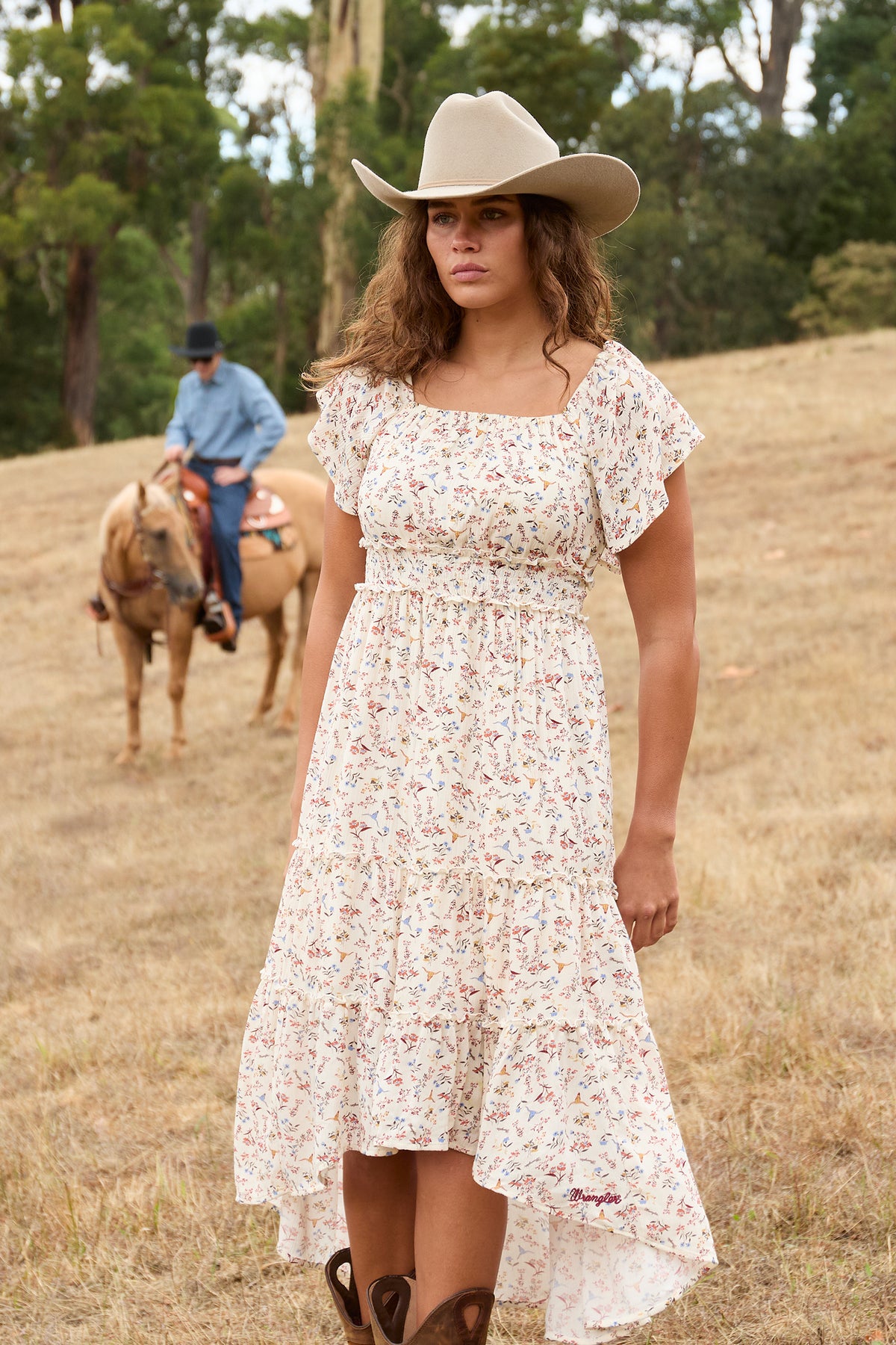 A woman wearing a short-sleeve, high-low floral dress and a beige cowboy hat stands in a dry grass field with trees in the background. A man on a horse is visible in the blurred background.