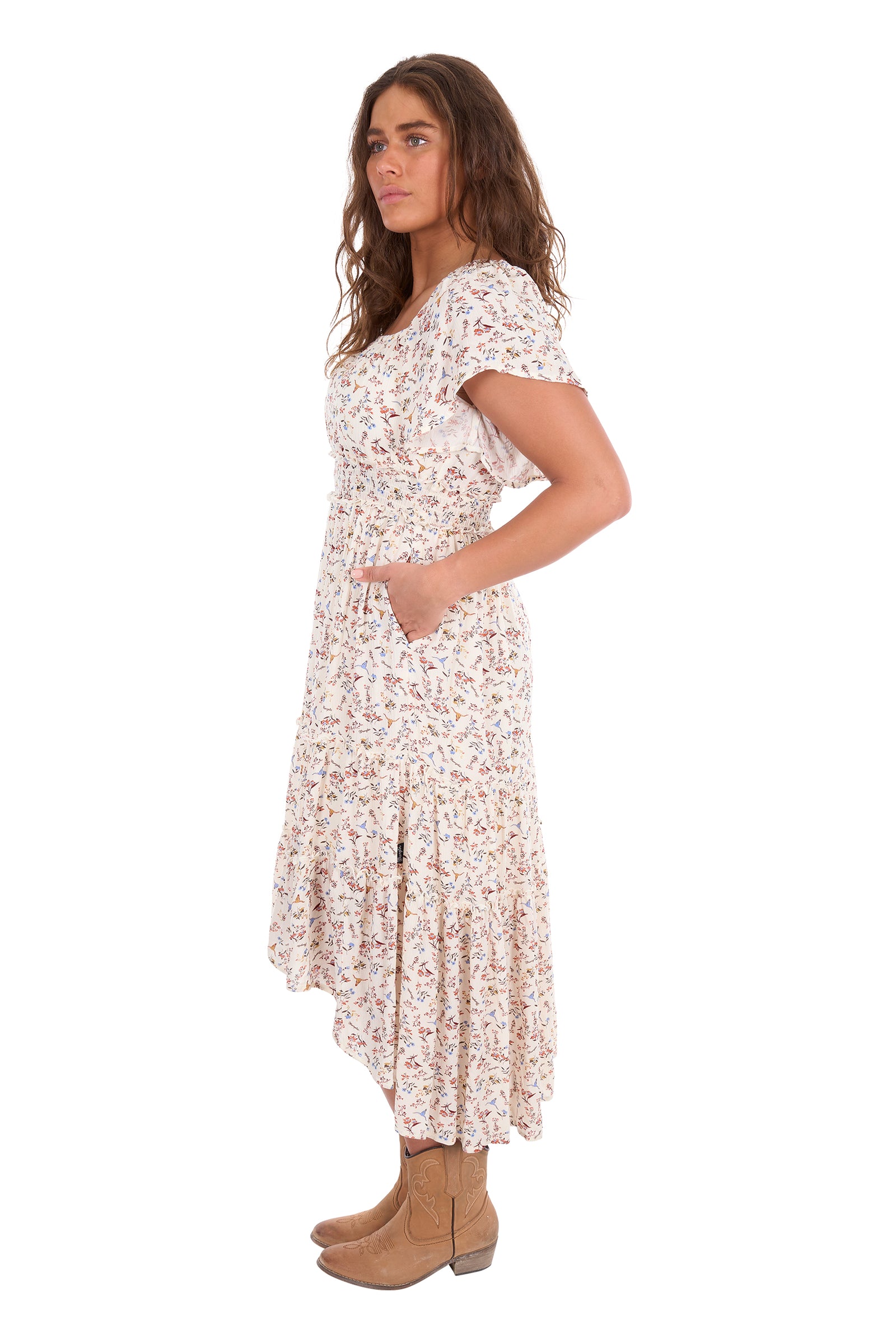 A woman modeling a short-sleeve midi dress with a delicate pink and green floral print on a cream background, paired with tan ankle boots, against a plain white studio background.