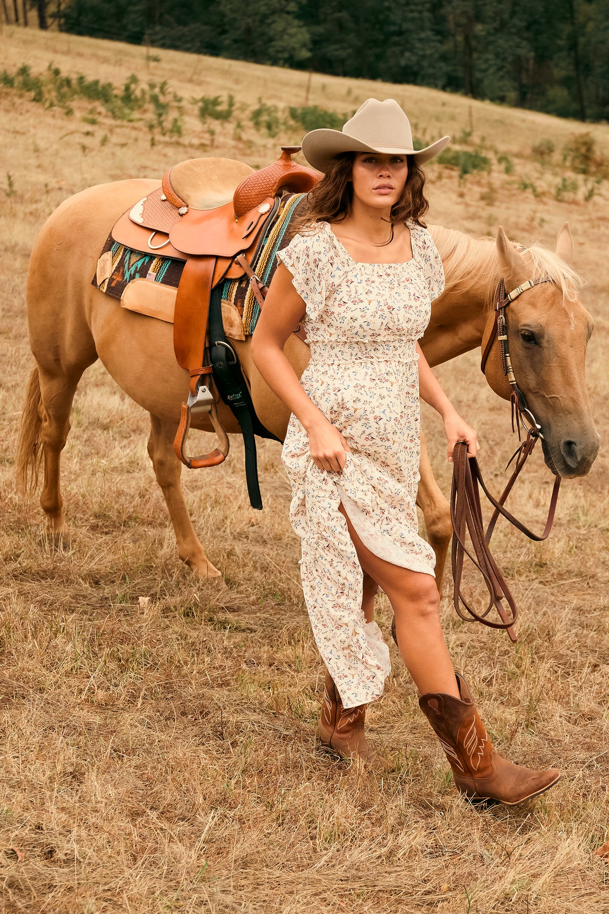 A woman in a white floral dress, cowboy hat, and brown cowboy boots walks alongside a light brown horse in a dry grass field. The horse has a brown leather saddle with a colorful patterned blanket.