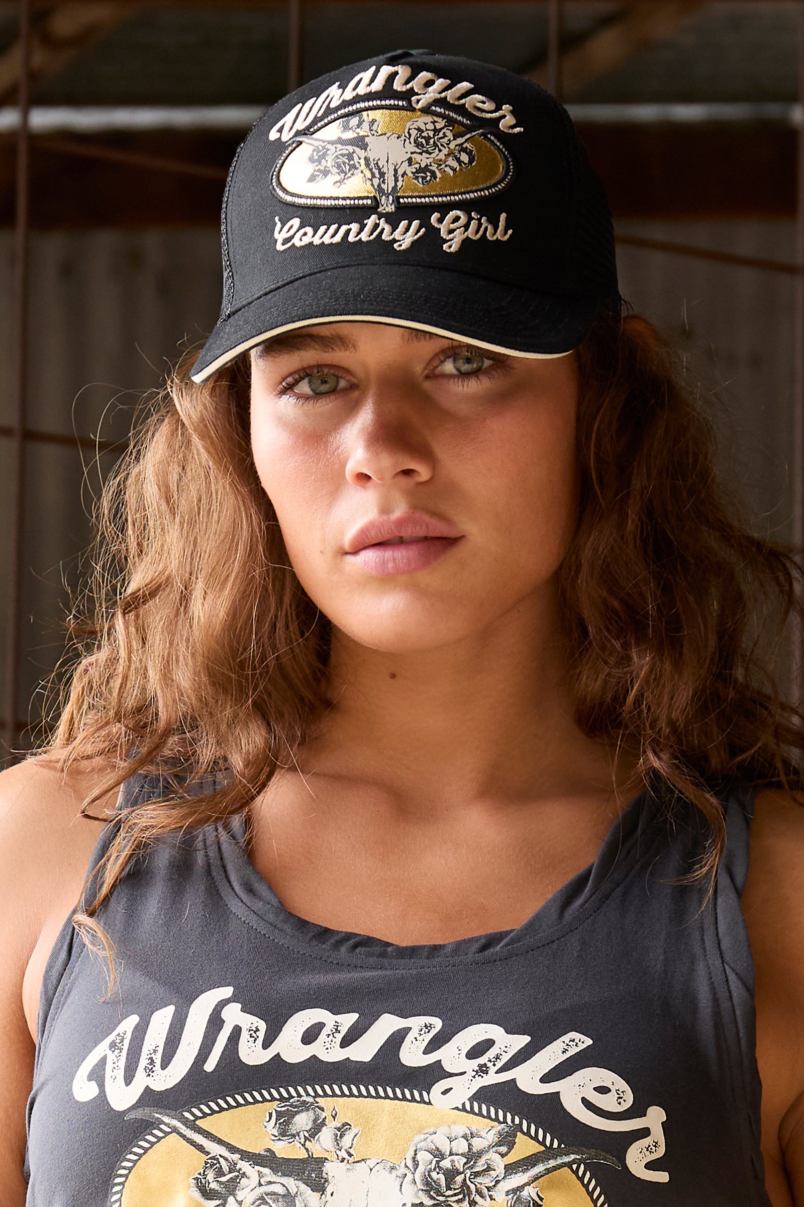 A woman wearing a black Wrangler baseball cap with a gold and white logo, paired with a dark gray tank top featuring a Wrangler graphic.