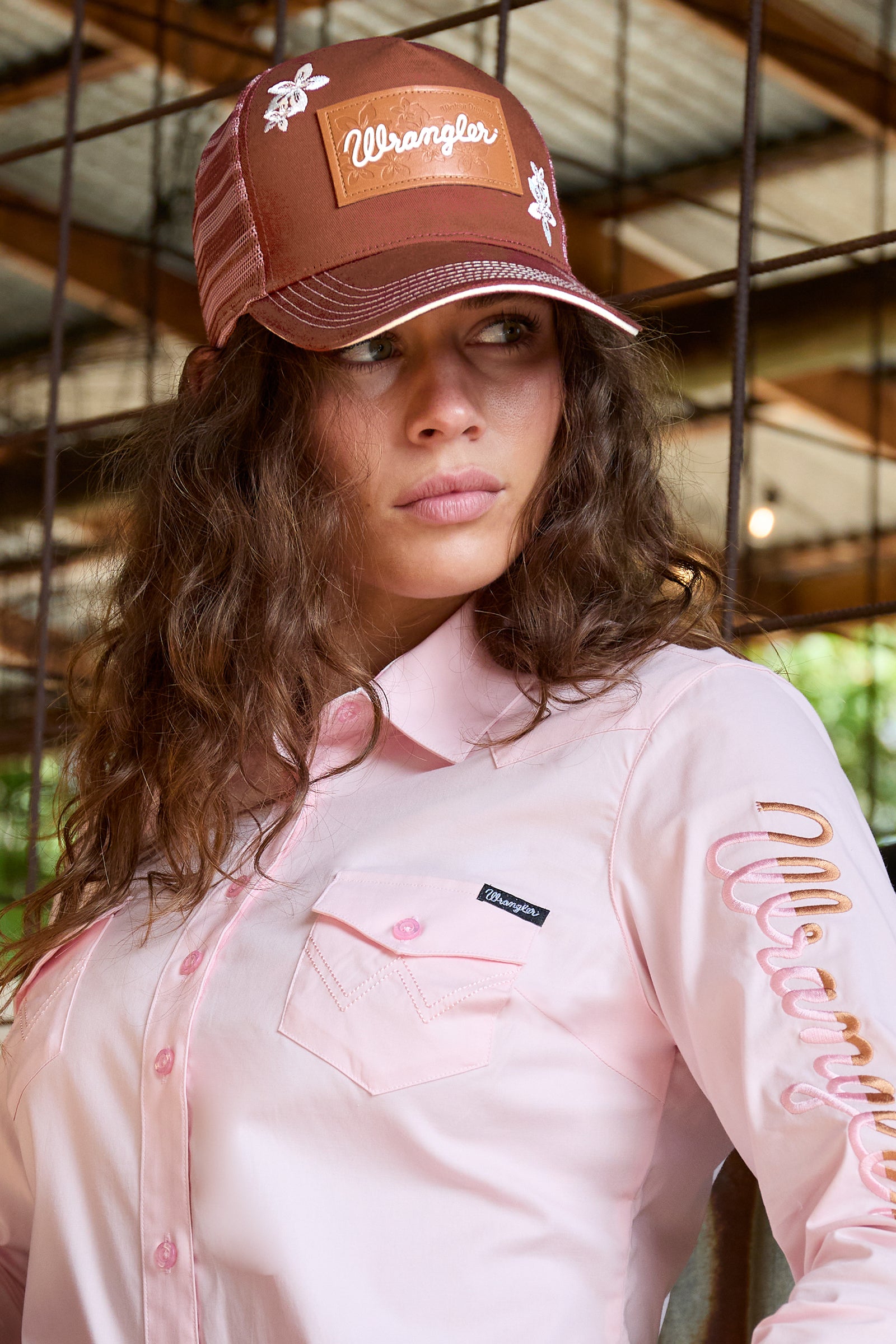 A woman wearing a brown Wrangler trucker hat and a light pink long-sleeve button-up shirt with embroidered details on the sleeve, posed in front of a rustic metal fence.