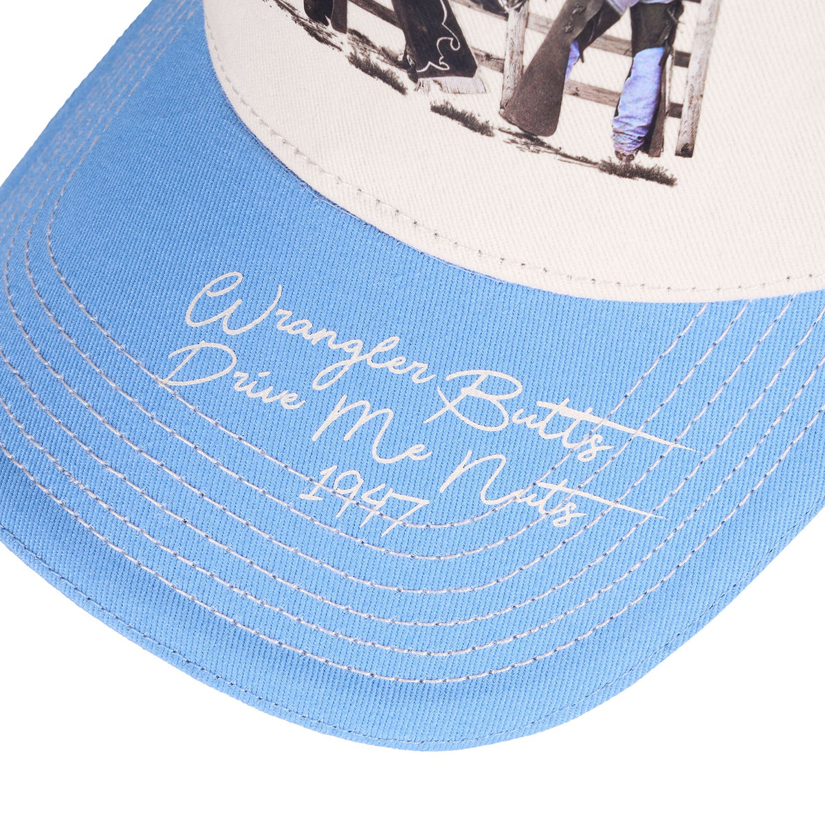 Close-up of a light blue and off-white baseball cap with white embroidered script reading 'Wrangler Butts Drink Me Nots 1947' on the brim.