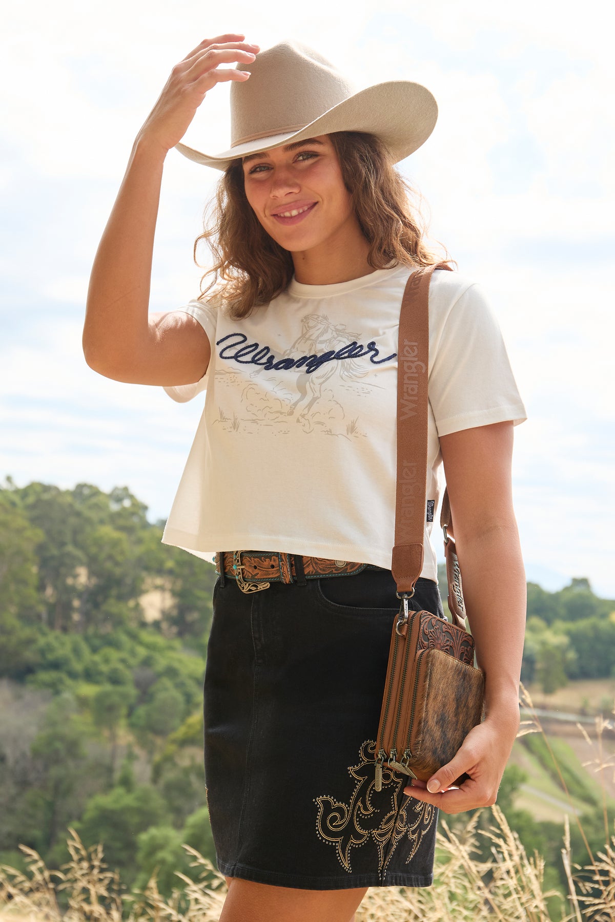 A woman outdoors wearing a cream western-style crop top with a dark script logo, a black denim skirt, a brown leather crossbody bag, and a beige cowboy hat.