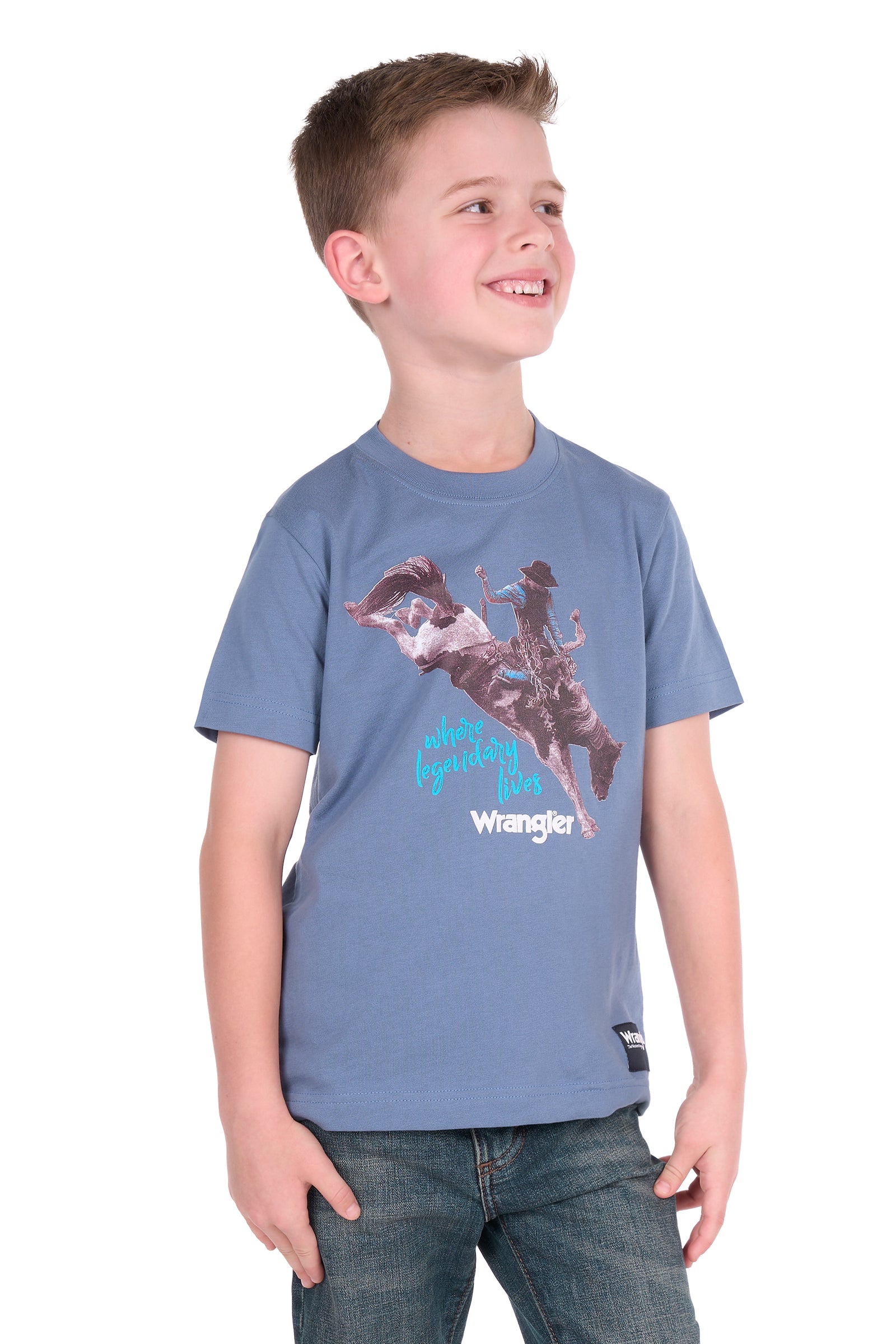 A young boy wearing a blue Wrangler t-shirt with a cowboy graphic and jeans, standing against a plain white background.