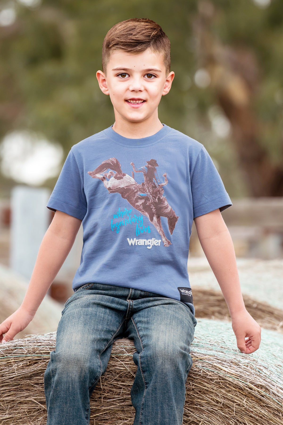 A young boy wearing a blue Wrangler t-shirt with a cowboy riding a bucking bronco graphic, sitting on a hay bale outdoors.