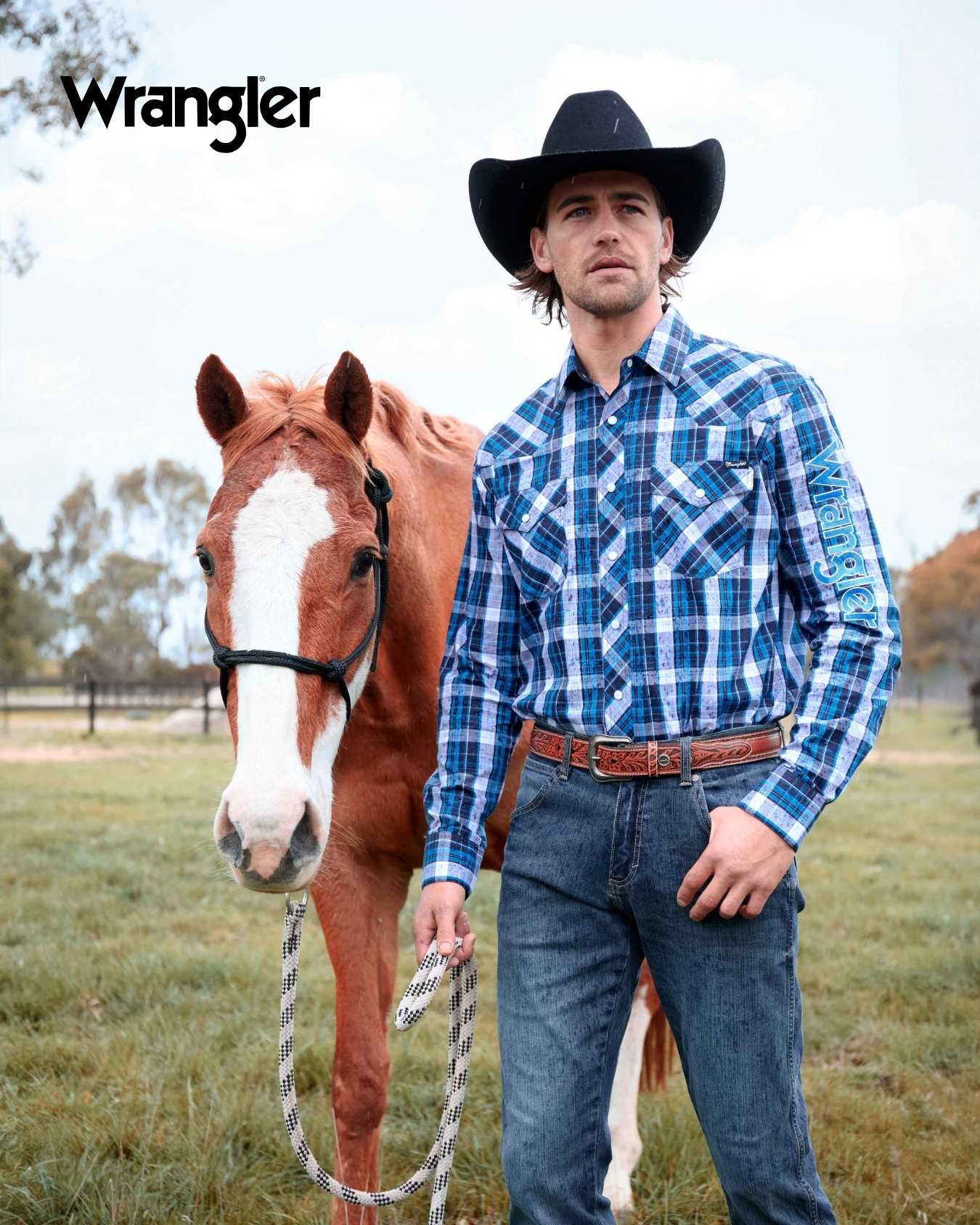 Man in blue plaid shirt and cowboy hat standing next to a horse in a field with Wrangler logo.