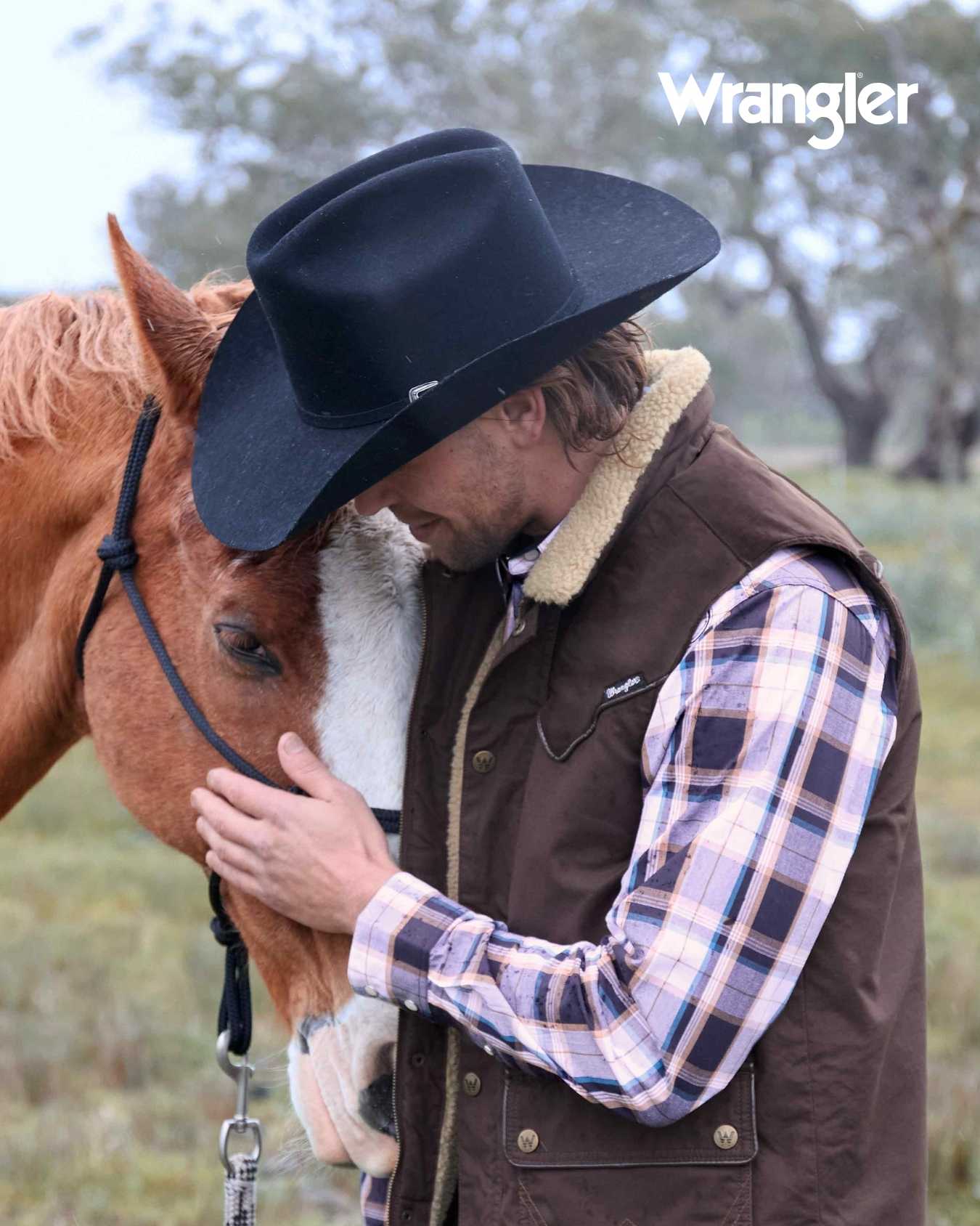 Man in cowboy hat and vest embracing a horse with 'Wrangler' branding.