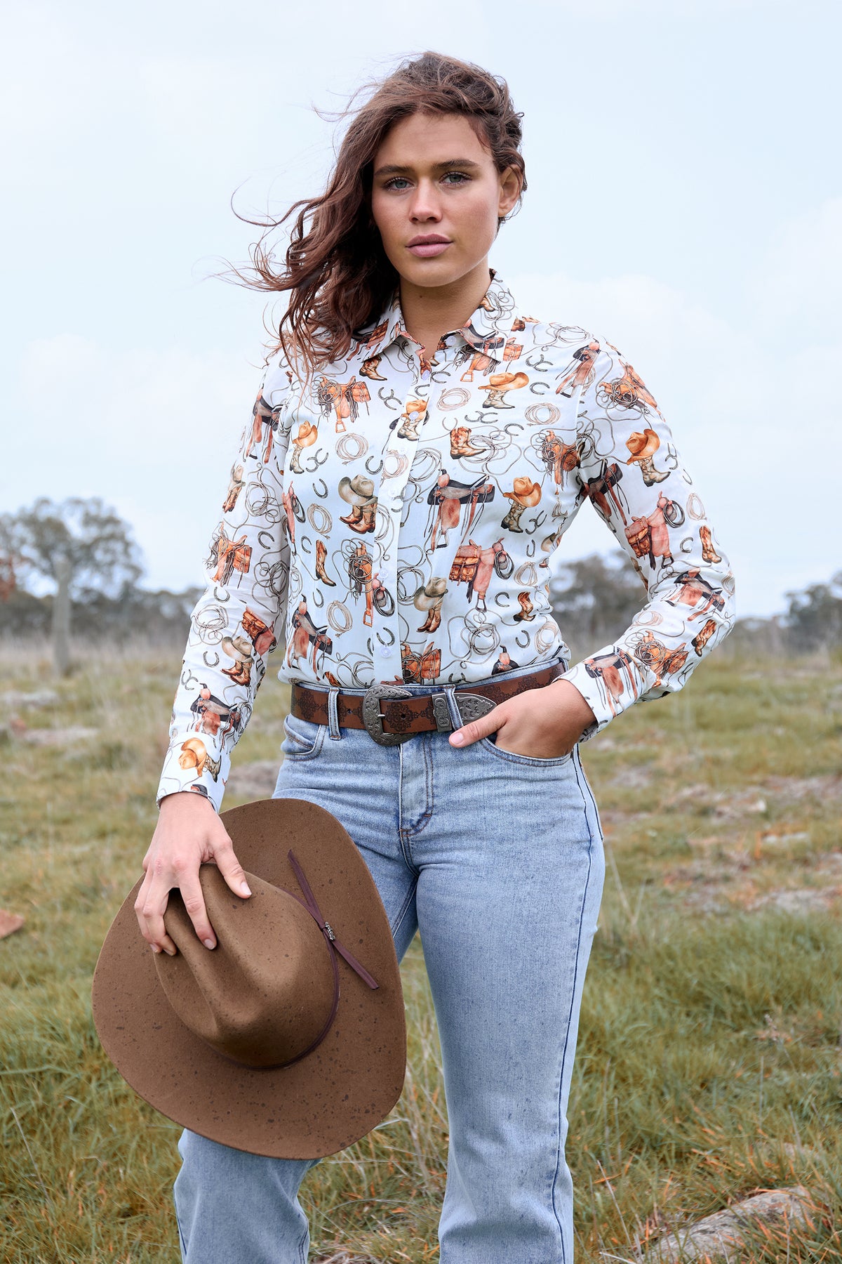 Woman wearing a patterned shirt and jeans, holding a hat in a grassy field.