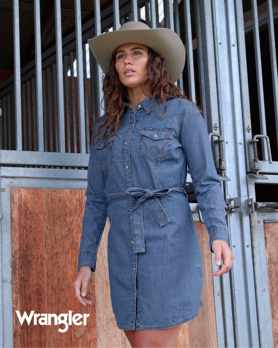 Woman wearing a blue denim dress and beige cowboy hat in front of a wooden door with 'Wrangler' branding.