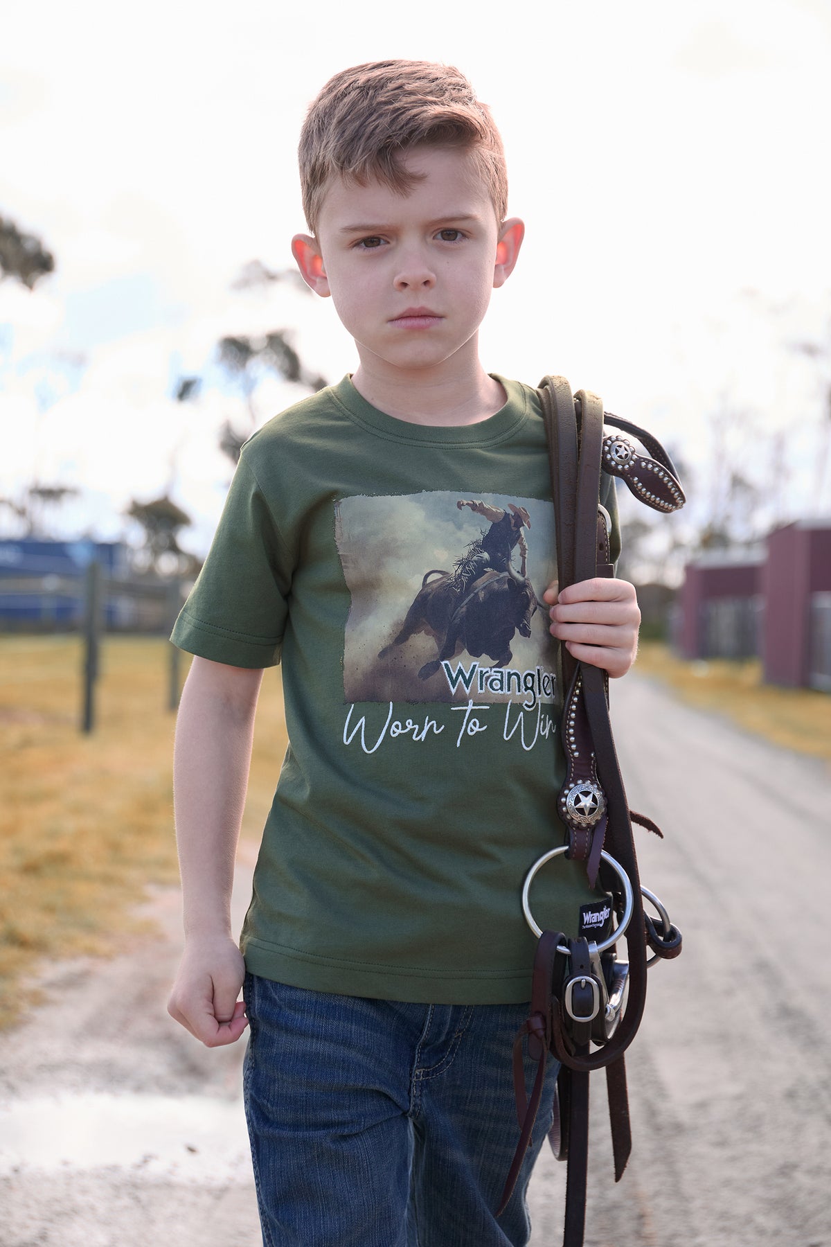 Young boy wearing a green t-shirt with a graphic design, holding a whip on a road.