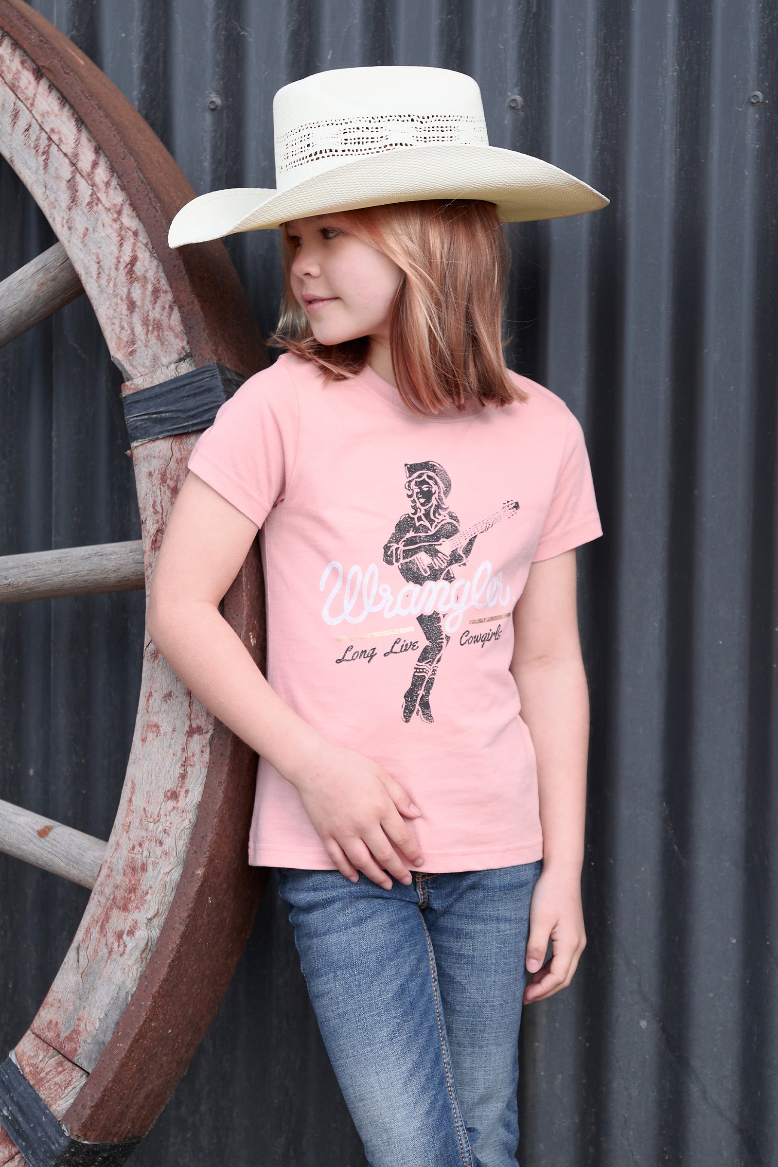 Child wearing a pink t-shirt with a graphic design and a cowboy hat, standing next to a large wheel.
