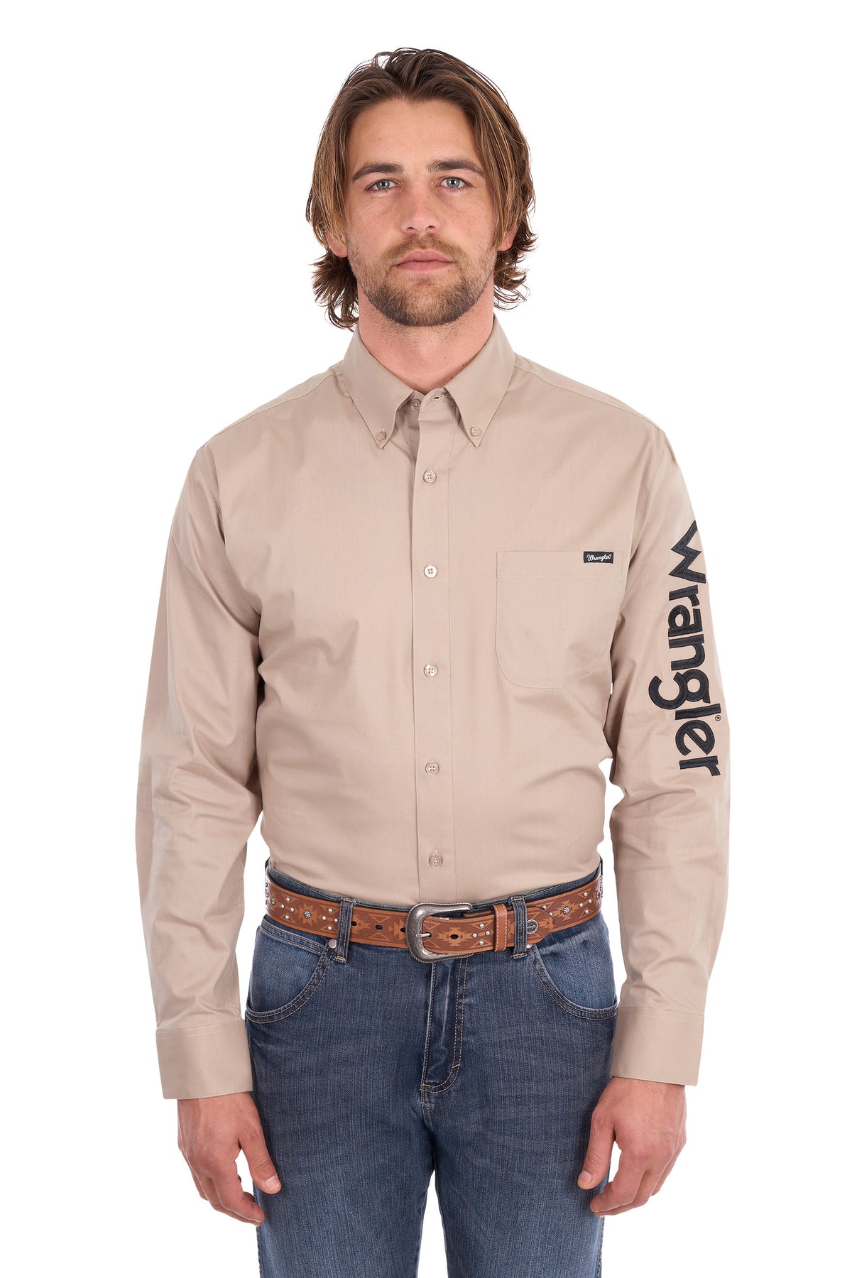A man wearing a beige Wrangler long-sleeve button-down shirt with a black logo printed vertically on the left sleeve, paired with blue jeans and a brown leather belt, against a plain white background.