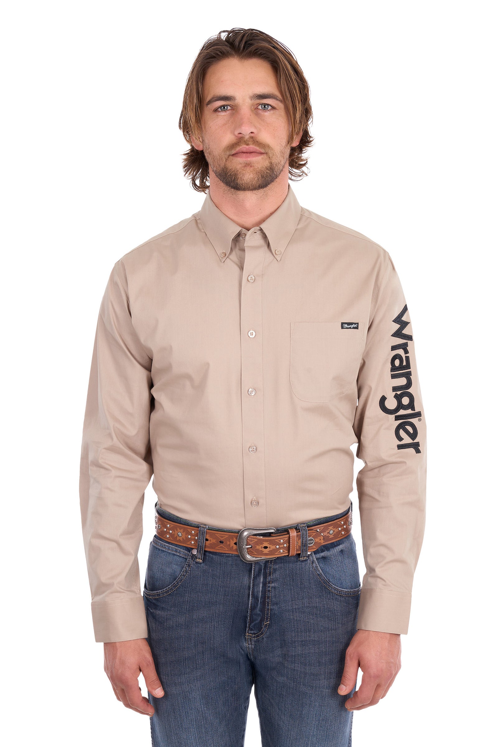 A man wearing a beige Wrangler long-sleeve button-down shirt with a black logo printed vertically on the left sleeve, paired with blue jeans and a brown leather belt, against a plain white background.