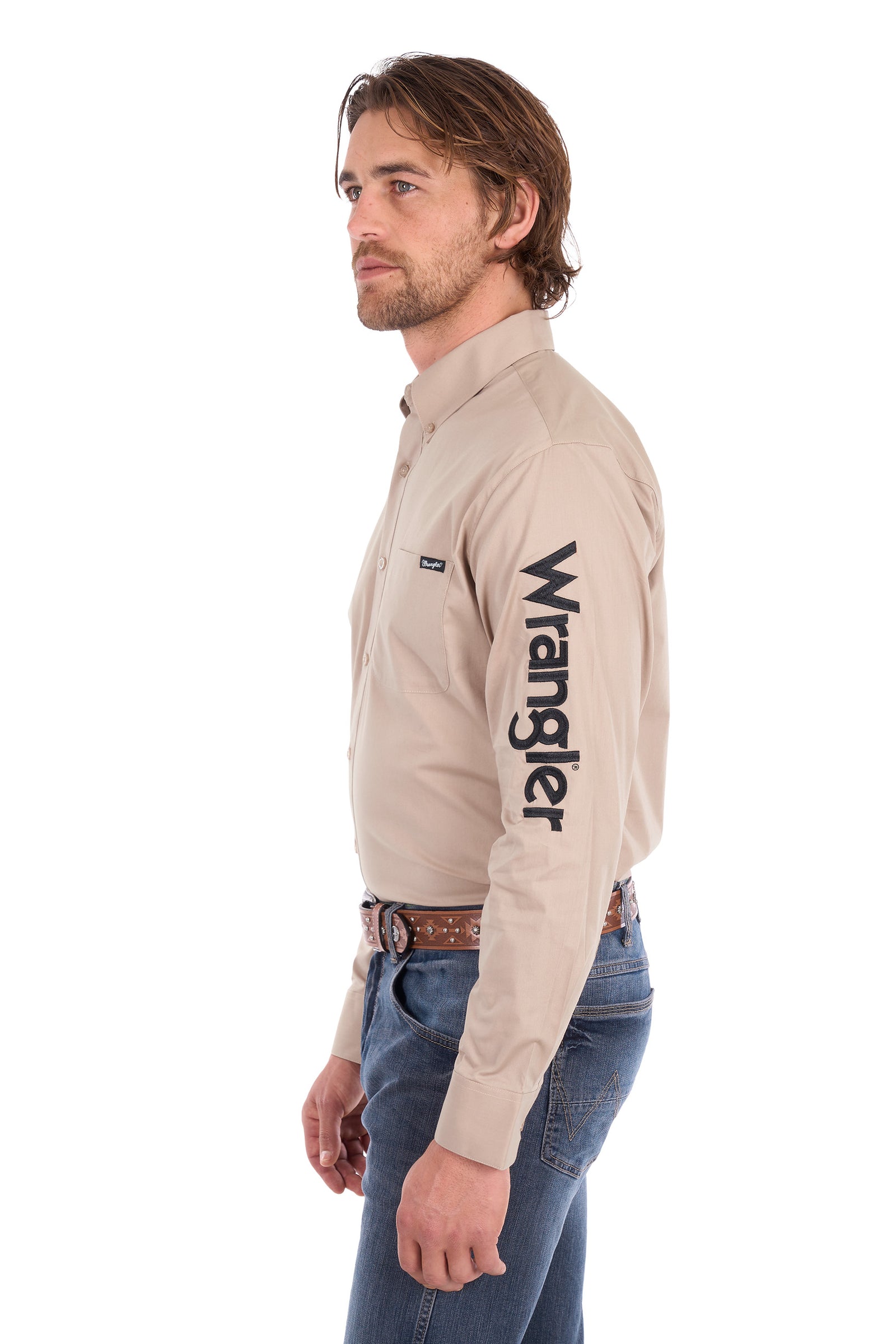 A man wearing a beige Wrangler long-sleeve button-down shirt with a black logo printed vertically on the left sleeve, paired with blue jeans, shown in profile against a white background.