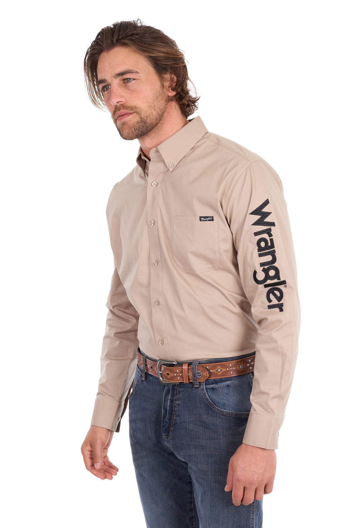 A man wearing a beige Wrangler long-sleeve button-down shirt with a black logo printed vertically on the left sleeve, paired with blue jeans and a brown leather belt, photographed against a solid white background.