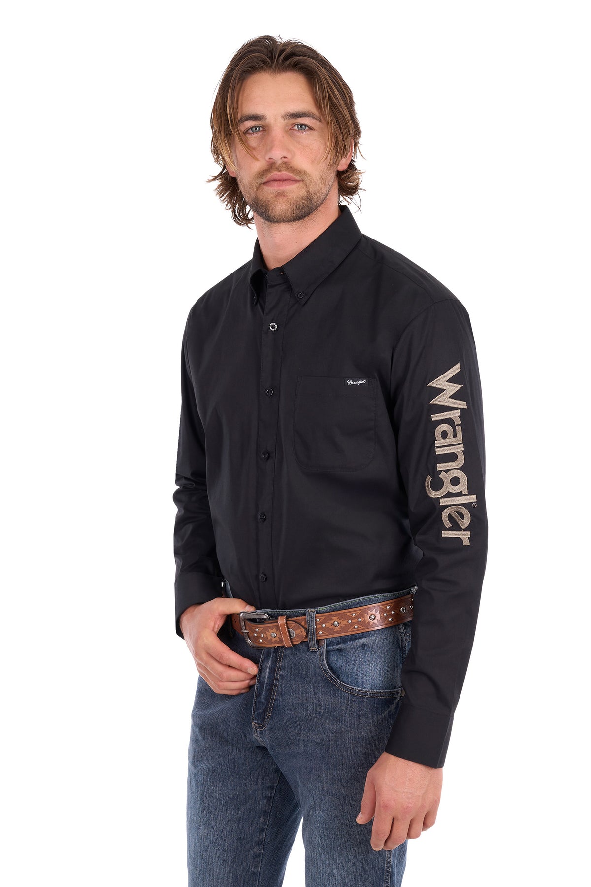 A man wearing a black Wrangler long-sleeve button-down shirt with a large beige logo printed vertically on the right sleeve, paired with blue jeans and a brown patterned belt, against a plain white background.
