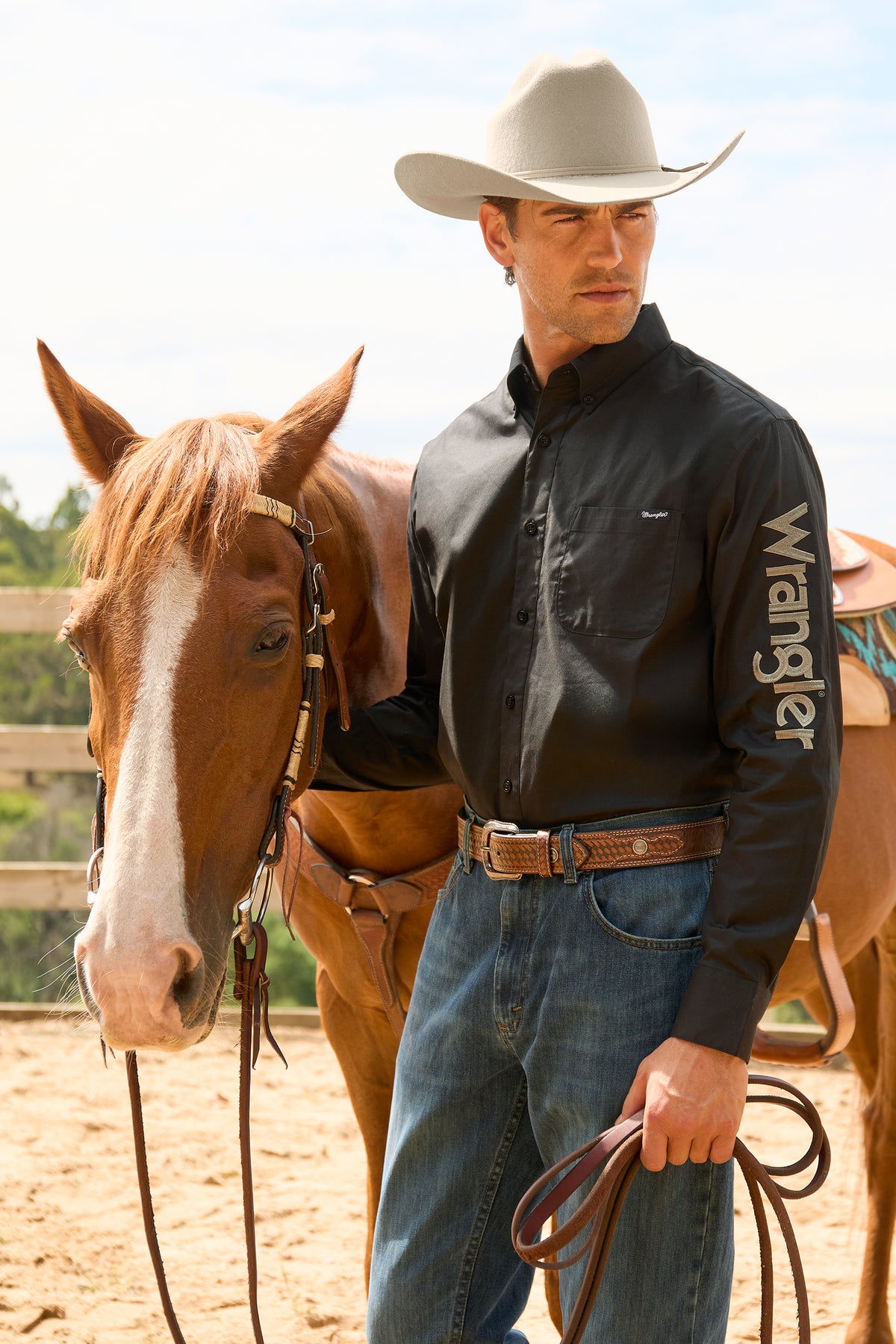 A man in a black long-sleeve Wrangler shirt and blue jeans stands beside a brown horse in an outdoor ranch setting, holding a leather lasso.