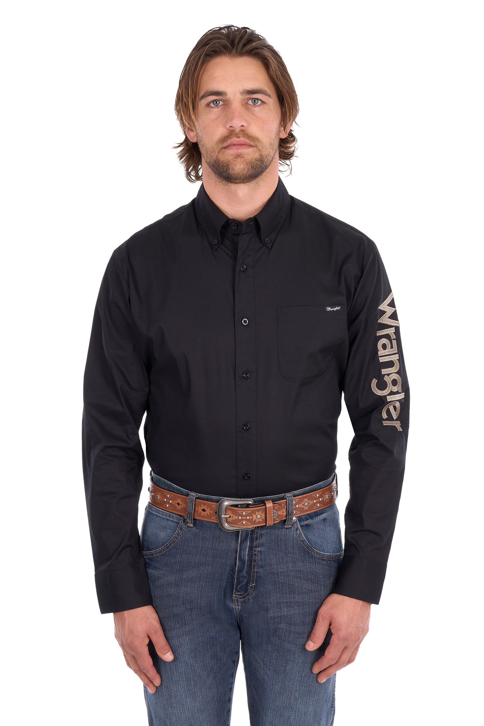 A man wearing a black long-sleeve button-down shirt with a large white 'Wrangler' logo printed vertically on the right sleeve, paired with blue jeans and a brown belt, against a plain white background.