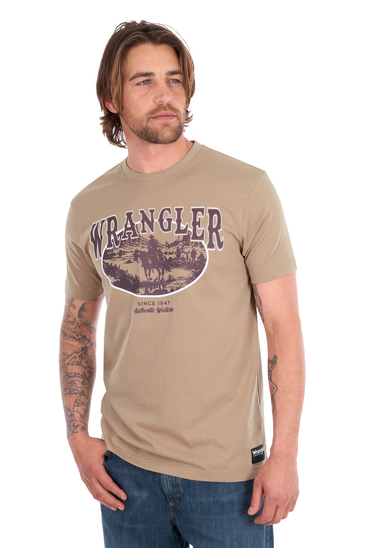 A man wearing a tan Wrangler short-sleeve t-shirt with a vintage-style western graphic on the chest, paired with blue jeans, against a plain white background.