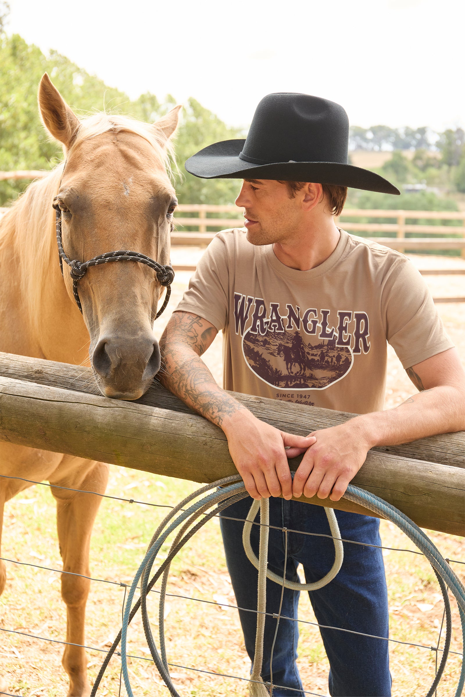 A man wearing a black cowboy hat and a beige Wrangler t-shirt leans on a wooden fence next to a light brown horse in an outdoor paddock.
