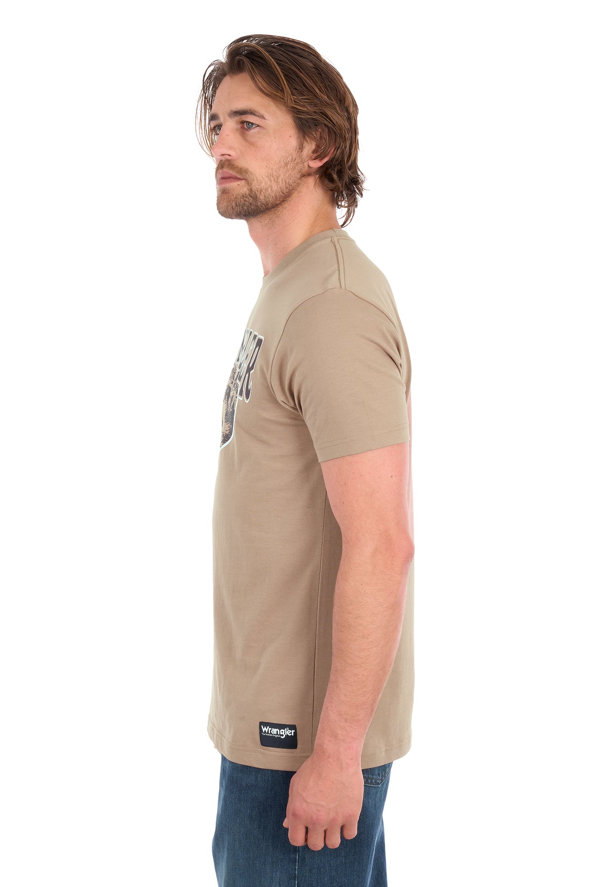 A man shown in profile wearing a light tan short-sleeve t-shirt with a graphic on the chest and a black Wrangler brand patch near the hem, paired with blue jeans against a white background.