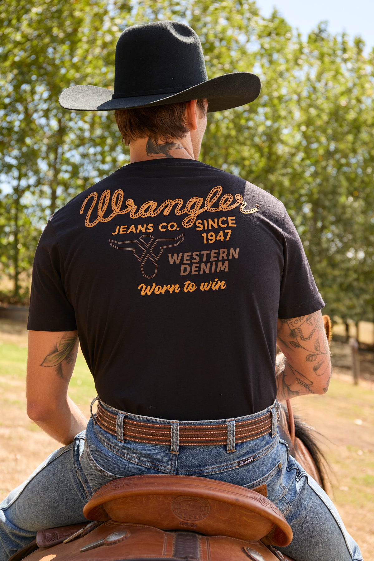 A man wearing a black Wrangler t-shirt from the back, sitting on a brown leather saddle outdoors. The shirt features a large tan and white graphic on the back reading 'Wrangler JEANS CO.