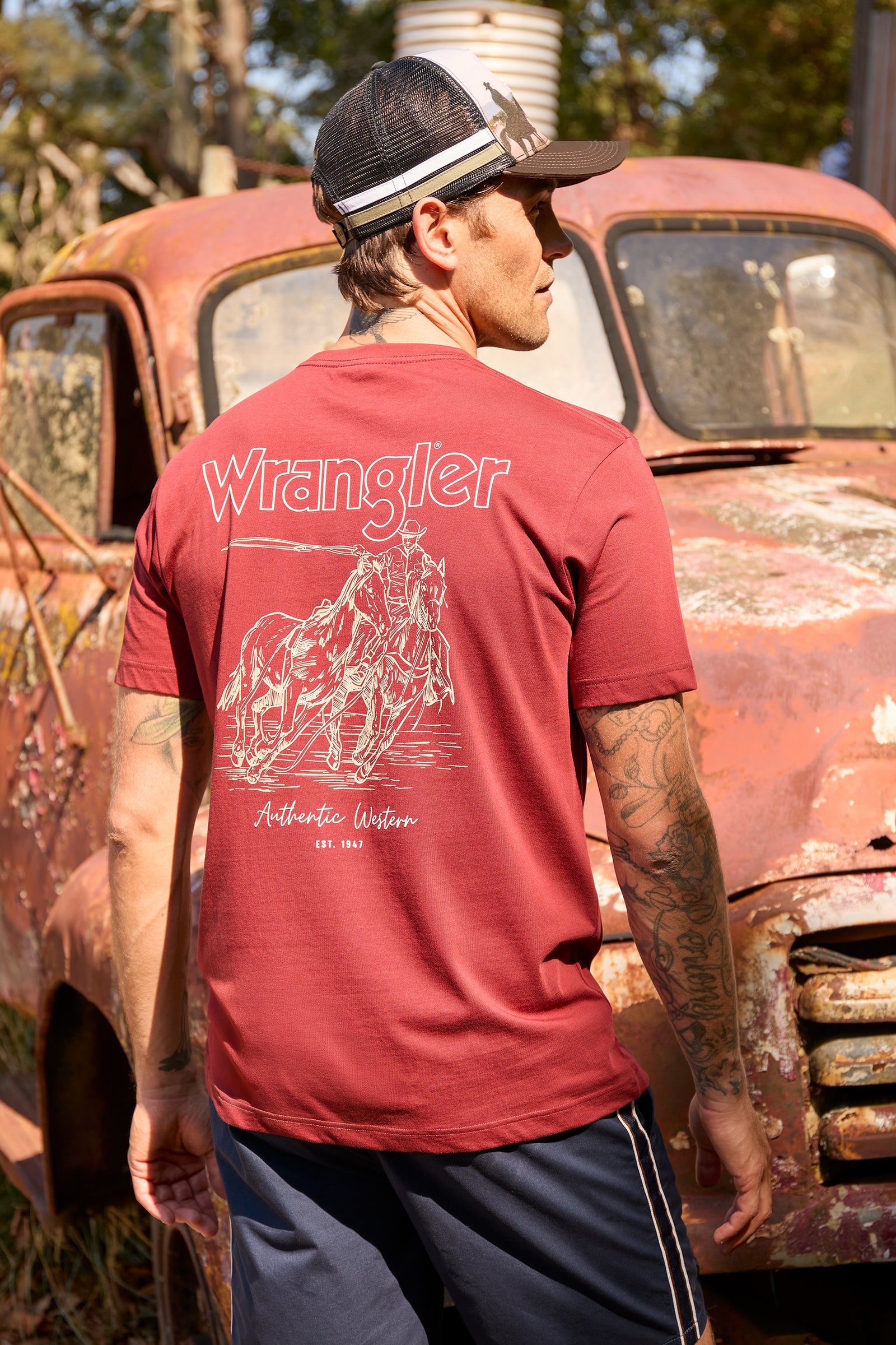 A man wearing a red Wrangler t-shirt with a white graphic print on the back, standing in front of a rusted vintage car outdoors.