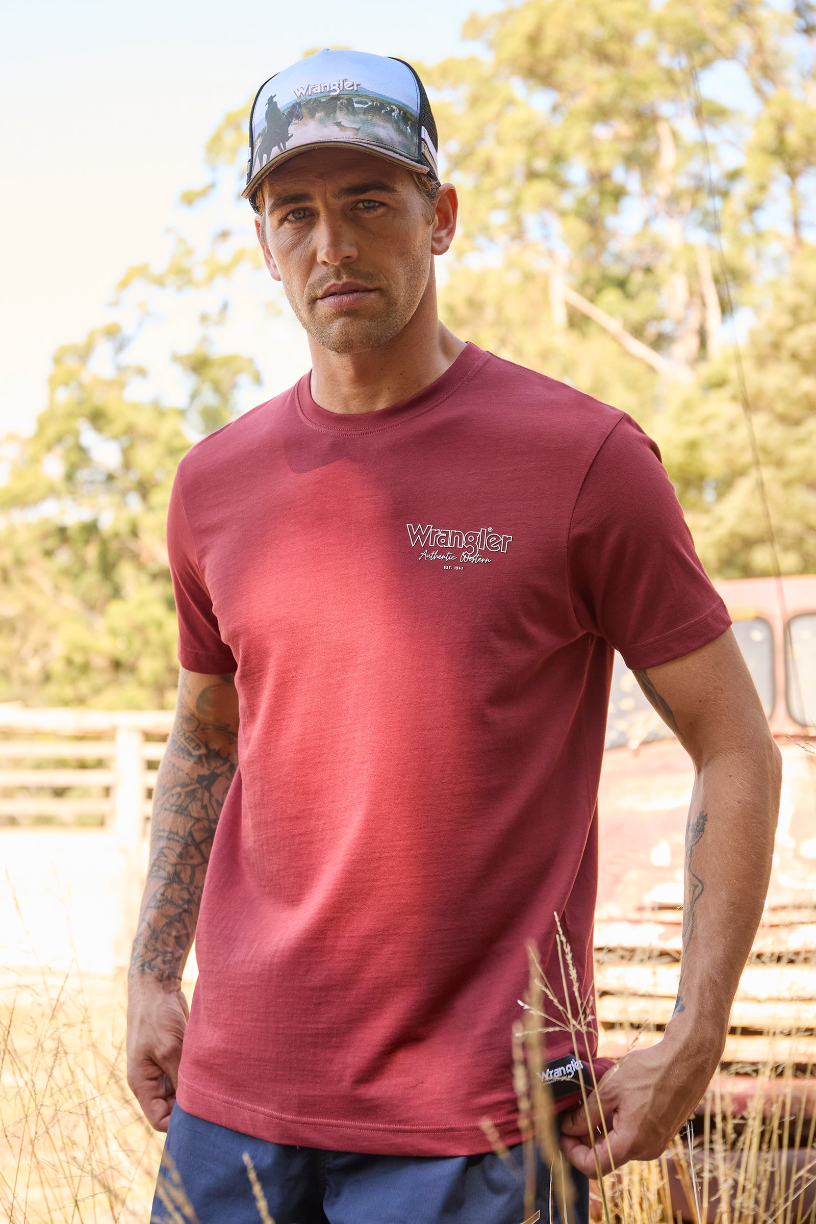 A man wearing a maroon Wrangler short-sleeve t-shirt and a black and white trucker hat, standing outdoors in a field with trees and a rustic vehicle in the background.