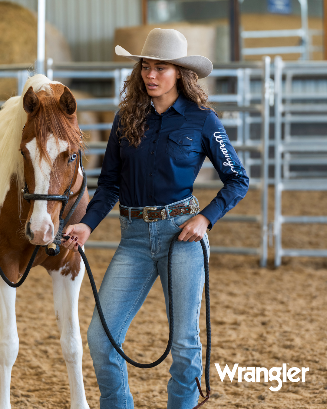 A woman in a cowboy hat and Wrangler navy blue long-sleeve shirt stands in a horse arena holding the reins of a brown and white paint horse. The Wrangler logo is visible in the bottom right corner.