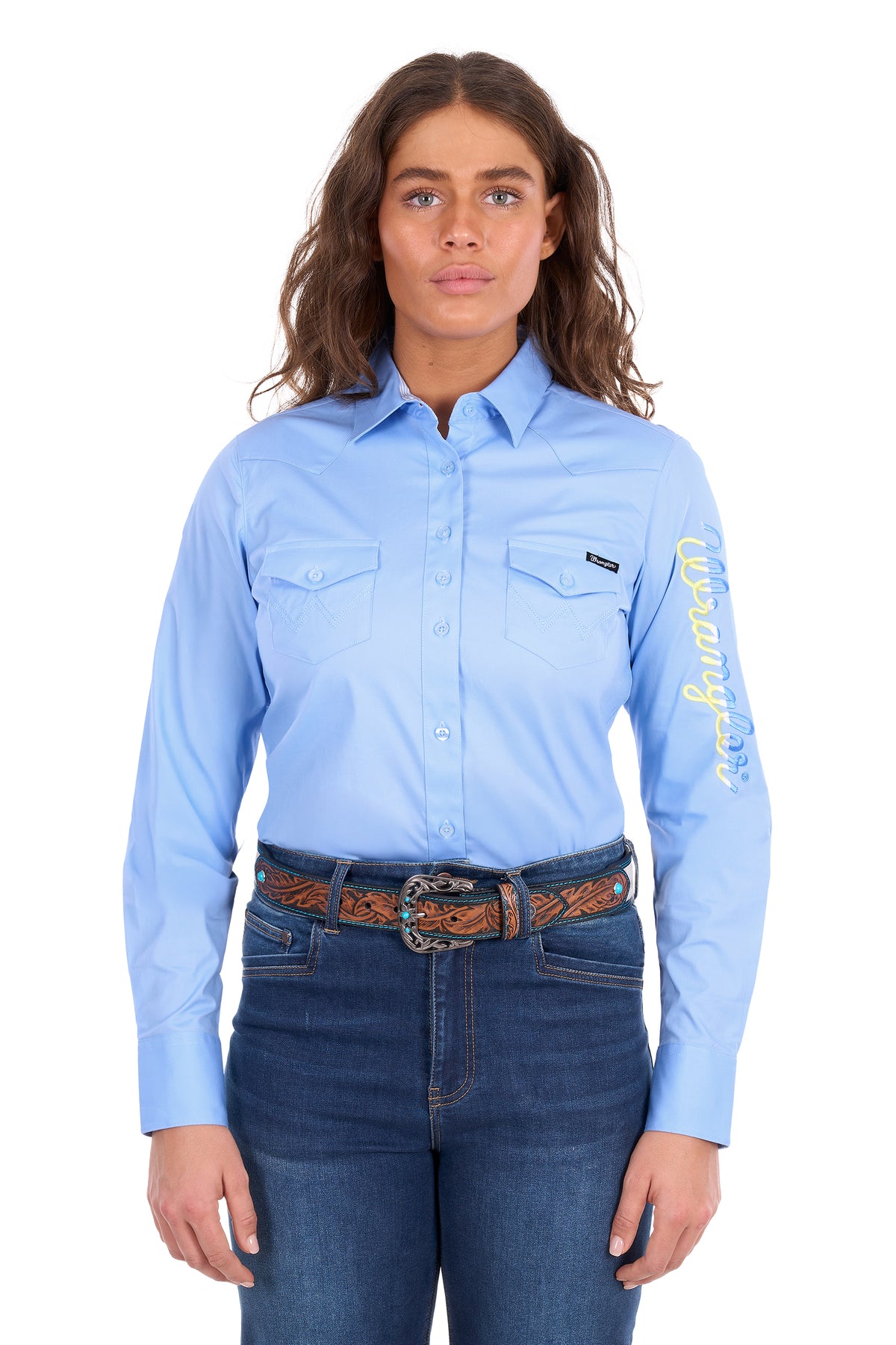 A woman modeling a light blue long-sleeve button-up shirt with two chest pockets, a black chest label, and yellow script text down one sleeve, paired with dark blue jeans and a brown patterned belt against a white background.