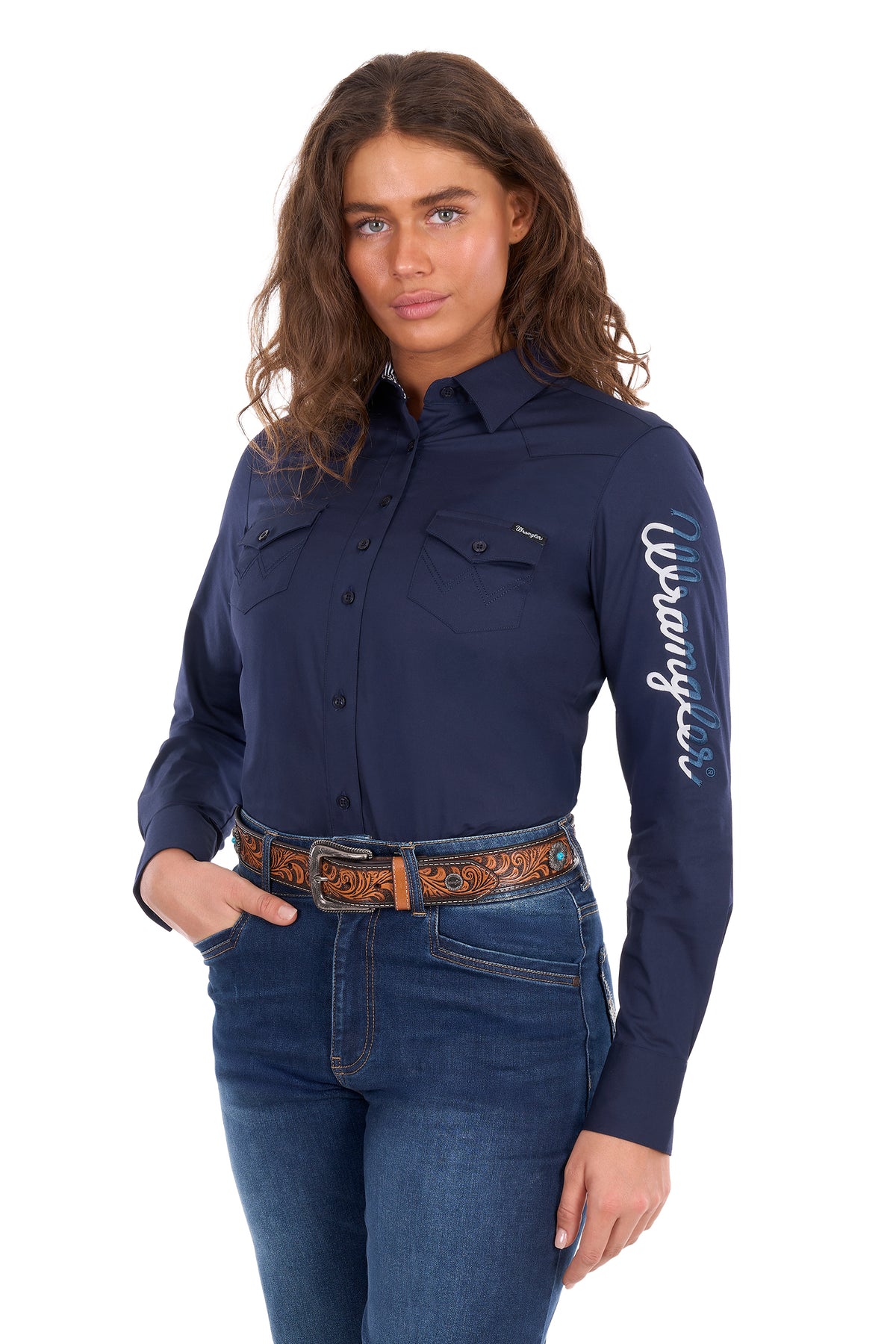 A woman modeling a navy blue long-sleeve button-up shirt with 'Wrangler' printed in white script down the right sleeve, paired with blue jeans and a brown western belt against a white background.