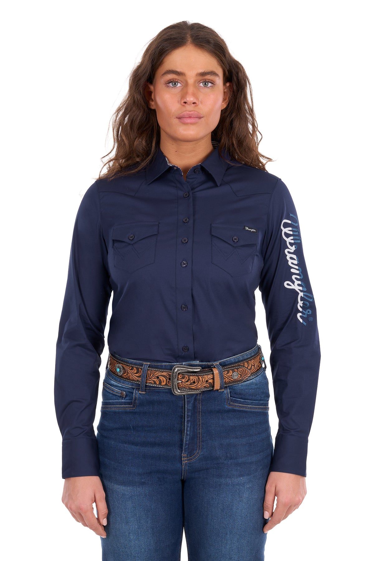 A woman modeling a navy blue long-sleeve button-up shirt with 'Kipsta' written in white script on the right sleeve, paired with blue jeans and a brown patterned belt against a plain white background.