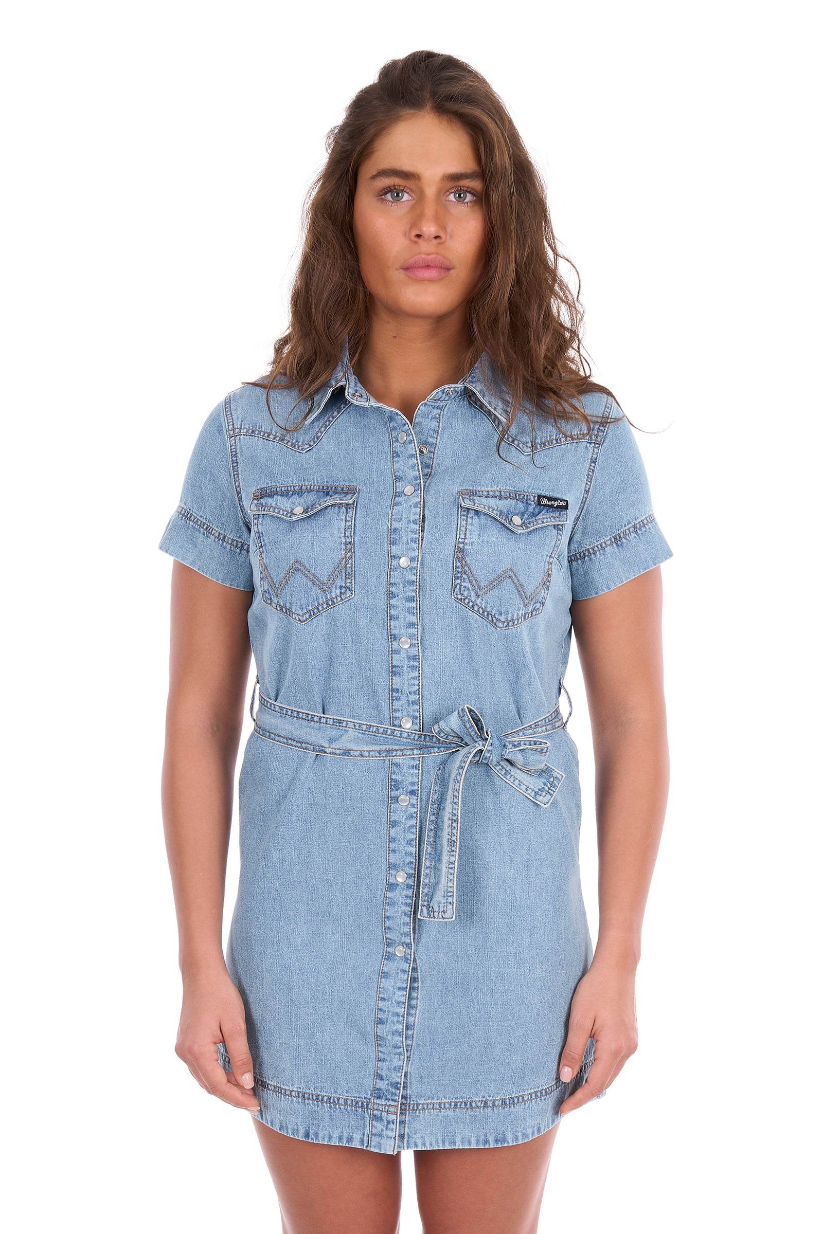 A woman modeling a light blue denim shirt dress with short sleeves, a button-front closure, two chest flap pockets, and a self-tie belt at the waist, photographed against a plain white background.