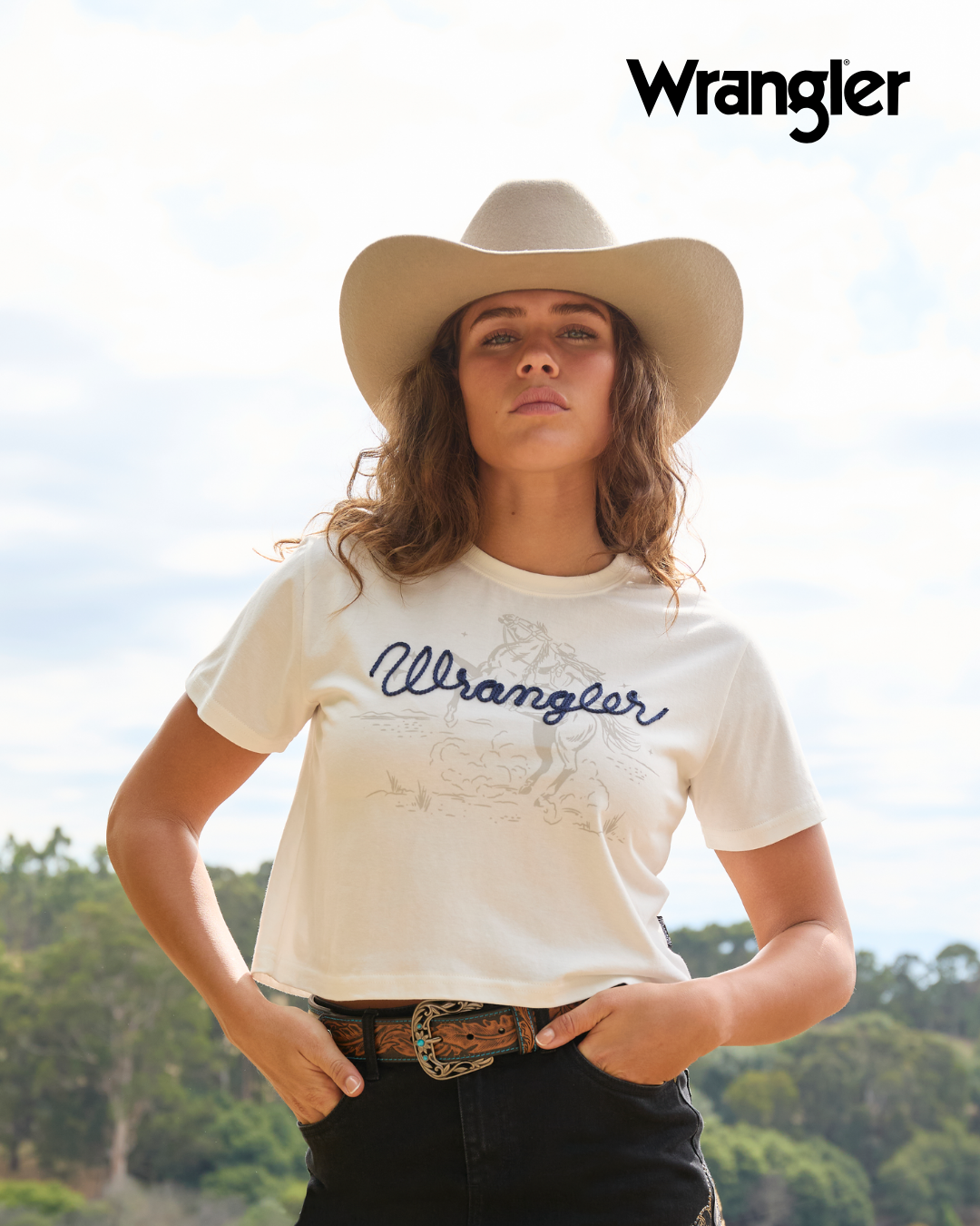 A woman wearing a beige cowboy hat and a cream Wrangler graphic t-shirt, paired with black jeans and a brown leather belt with a large buckle, set against an outdoor background with trees and a cloudy sky. The Wrangler logo is visible in the top right corner.
